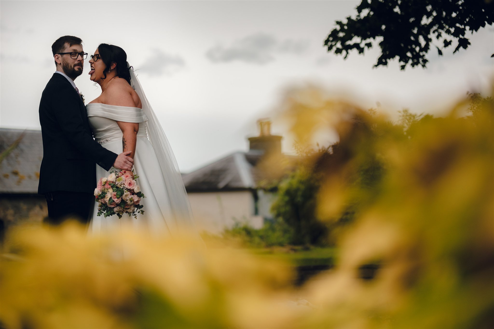 A newlywed couple at their wedding celebration, with the woman wearing an off-the-shoulder dress and holding a bouquet, and the man in a tuxedo, standing outdoors with a cloudy sky and rooftops in the background.