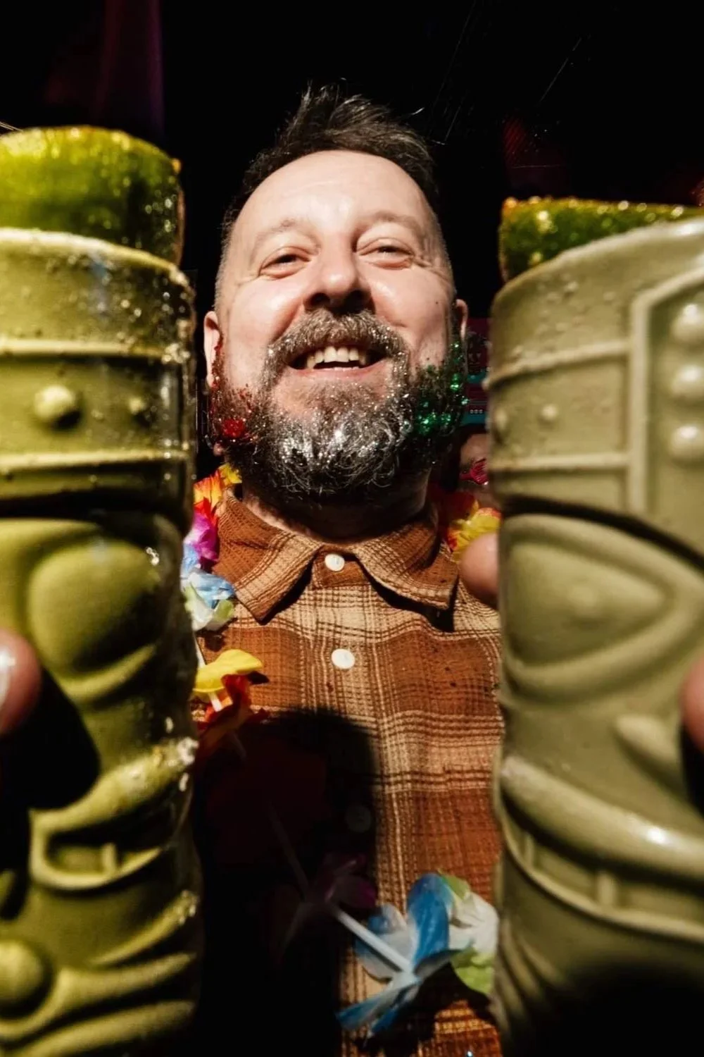 A joyful man with a beard holding colorful tiki mugs, smiling at a celebration or party.