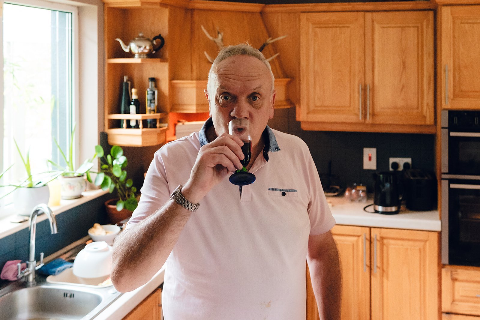 An elderly man in a pink polo shirt drinking a glass of dark soda in a kitchen with wooden cabinets, a countertop, and potted plants.
