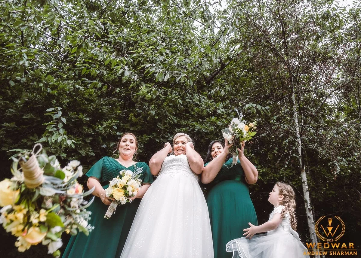 A bride and three women, possibly bridesmaids, standing outdoors under trees. The bride wears a white wedding dress and has a surprised or excited expression. The women wear dark green dresses and hold bouquets of flowers, one woman also holds a bouq