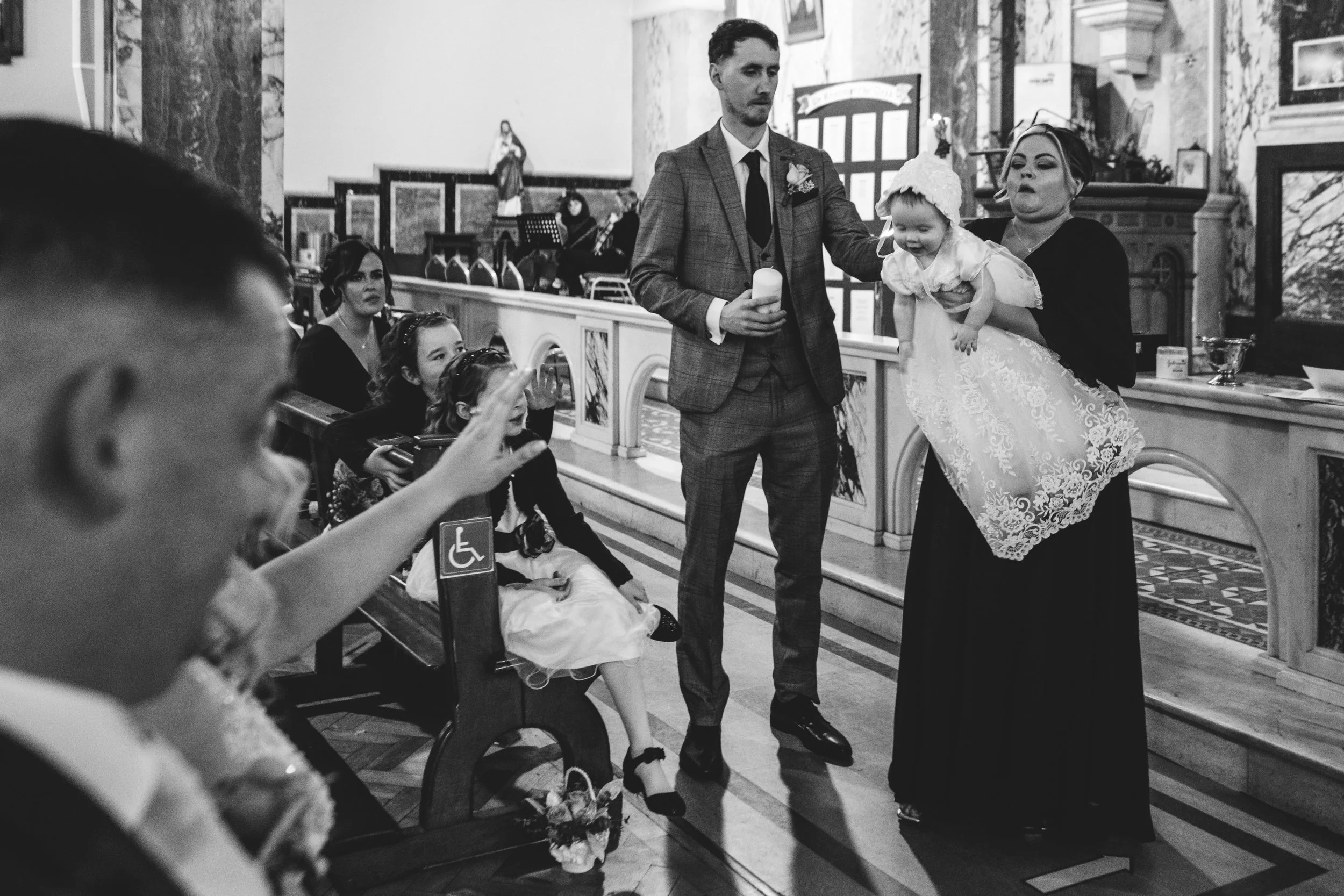 A black and white photo of a wedding ceremony inside a church, with a man in a suit holding a baby girl dressed in white, and a woman in dark clothing supporting the girl. Guests, including children, are seated nearby, with one person at the front ra