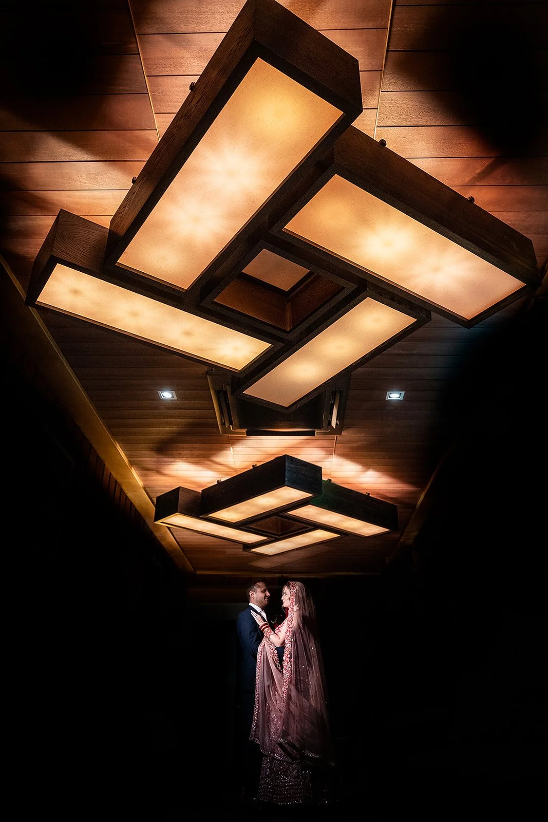 A bride and groom standing together under a modern, illuminated ceiling in a dark room, sharing a moment.