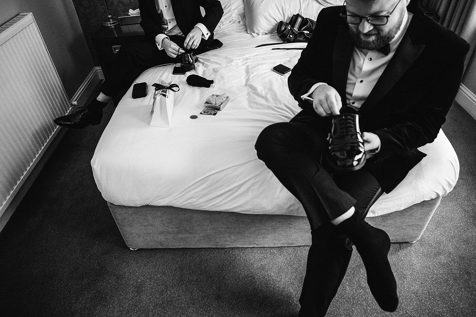 Two men in tuxedos are sitting on a bed in a hotel room. One man is adjusting his shoe, and the other is holding his shoe while sitting with his legs crossed. The bed has various small items on it, including a camera, a wallet, a phone, and some othe