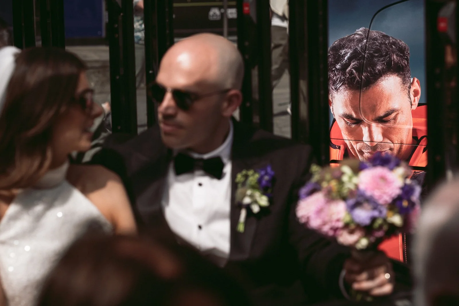 A man in a tuxedo and a boutonniere is seated next to a woman in a white gown, both smiling and talking. A person in an orange jacket is shown in a mirror or reflection behind them, holding a bouquet of flowers.
