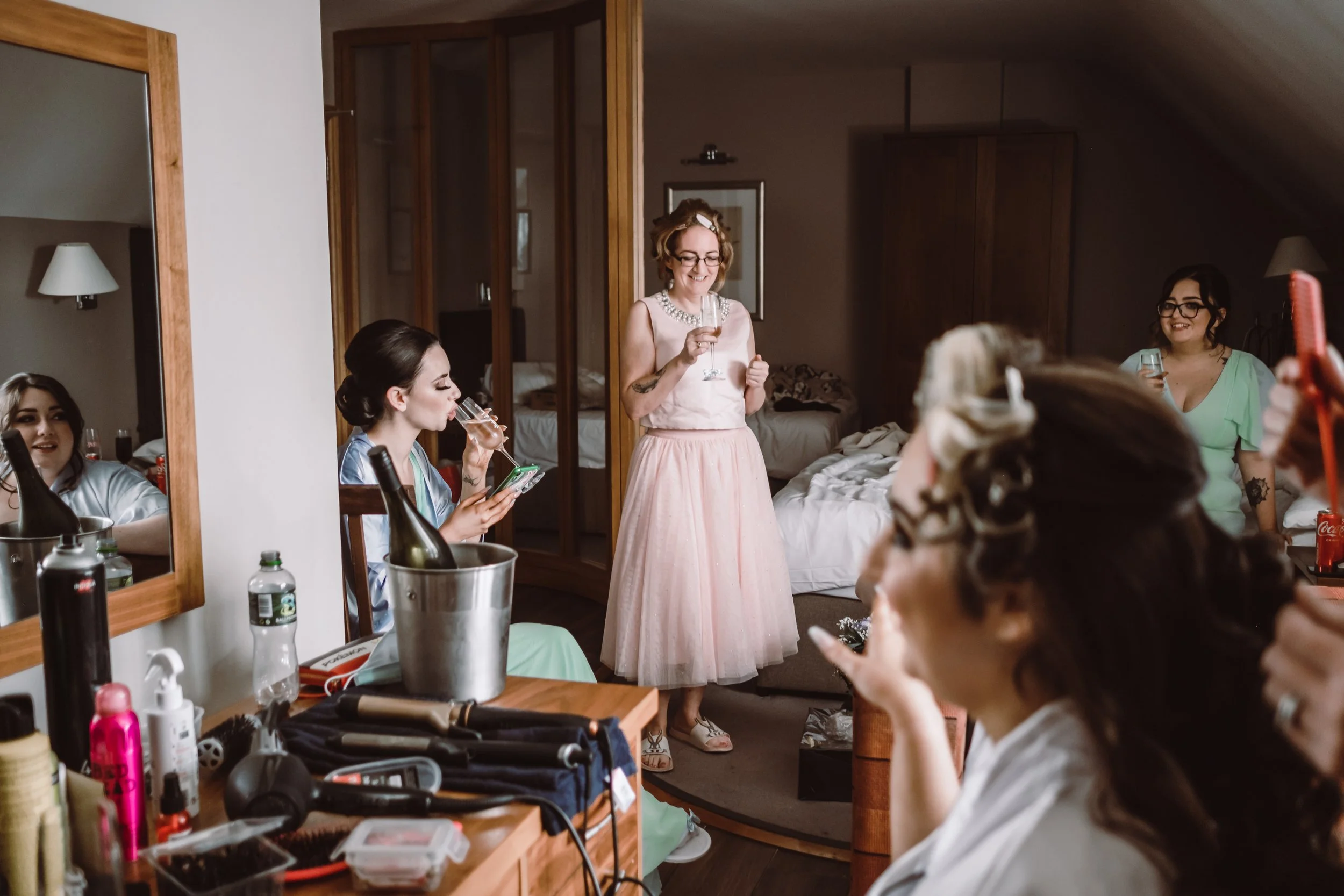 A group of women in a room getting ready, with makeup and hair tools on a vanity. One woman is sitting, looking in the mirror and holding a phone and drink, while another stands in a pink dress holding a drink and smiling. The room has mirrors and a 
