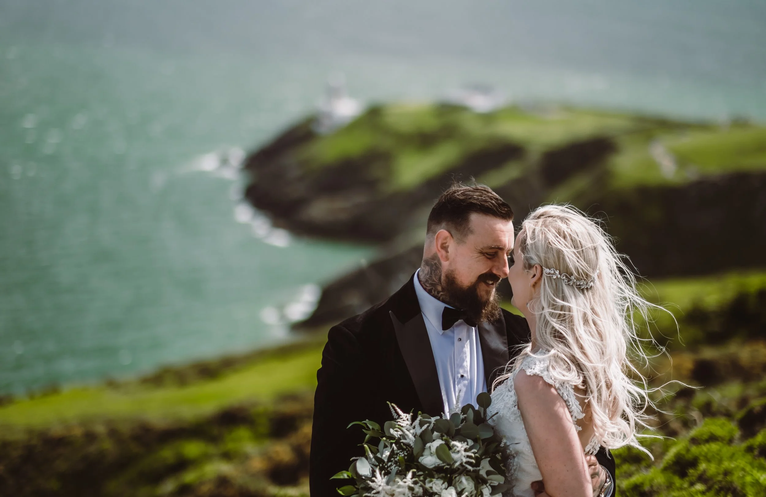 A bride and groom dressed in wedding attire sharing an intimate moment outdoors near the ocean, with rocky cliffs and green grass in the background.