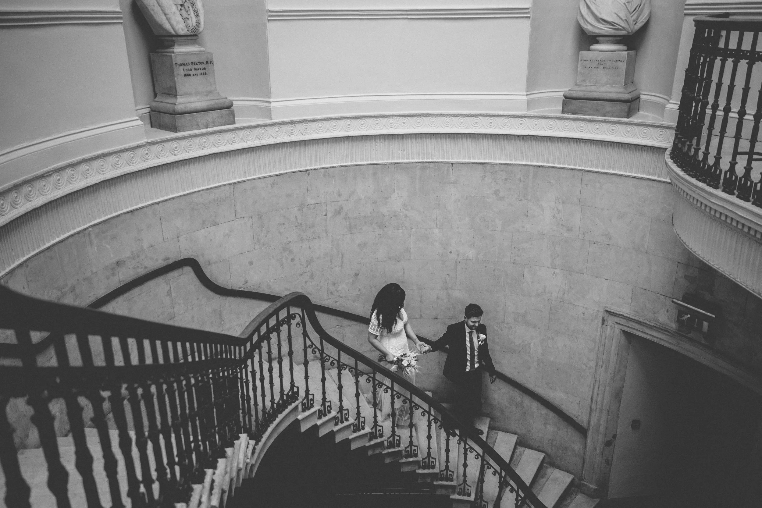A woman in a white dress and a man in a suit holding hands on a winding staircase with ornate railings, inside a building with decorative moldings and pedestal columns.