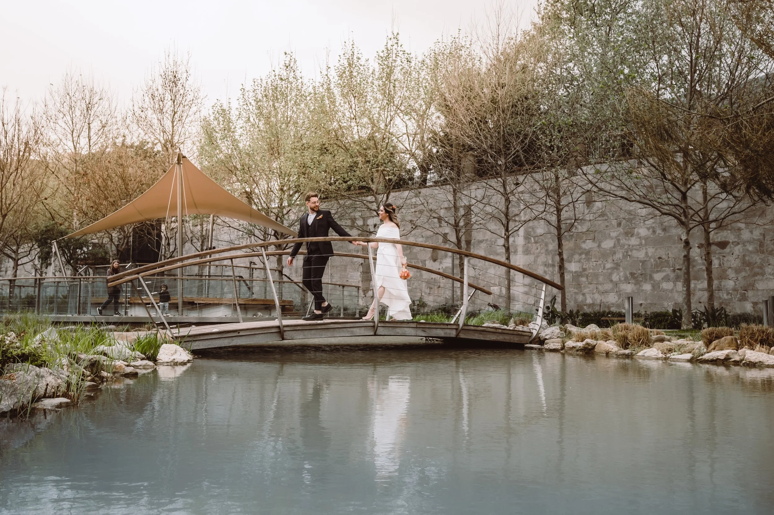 A bride and groom walking hand in hand across a small bridge over a pond, with bare trees and a stone wall in the background.