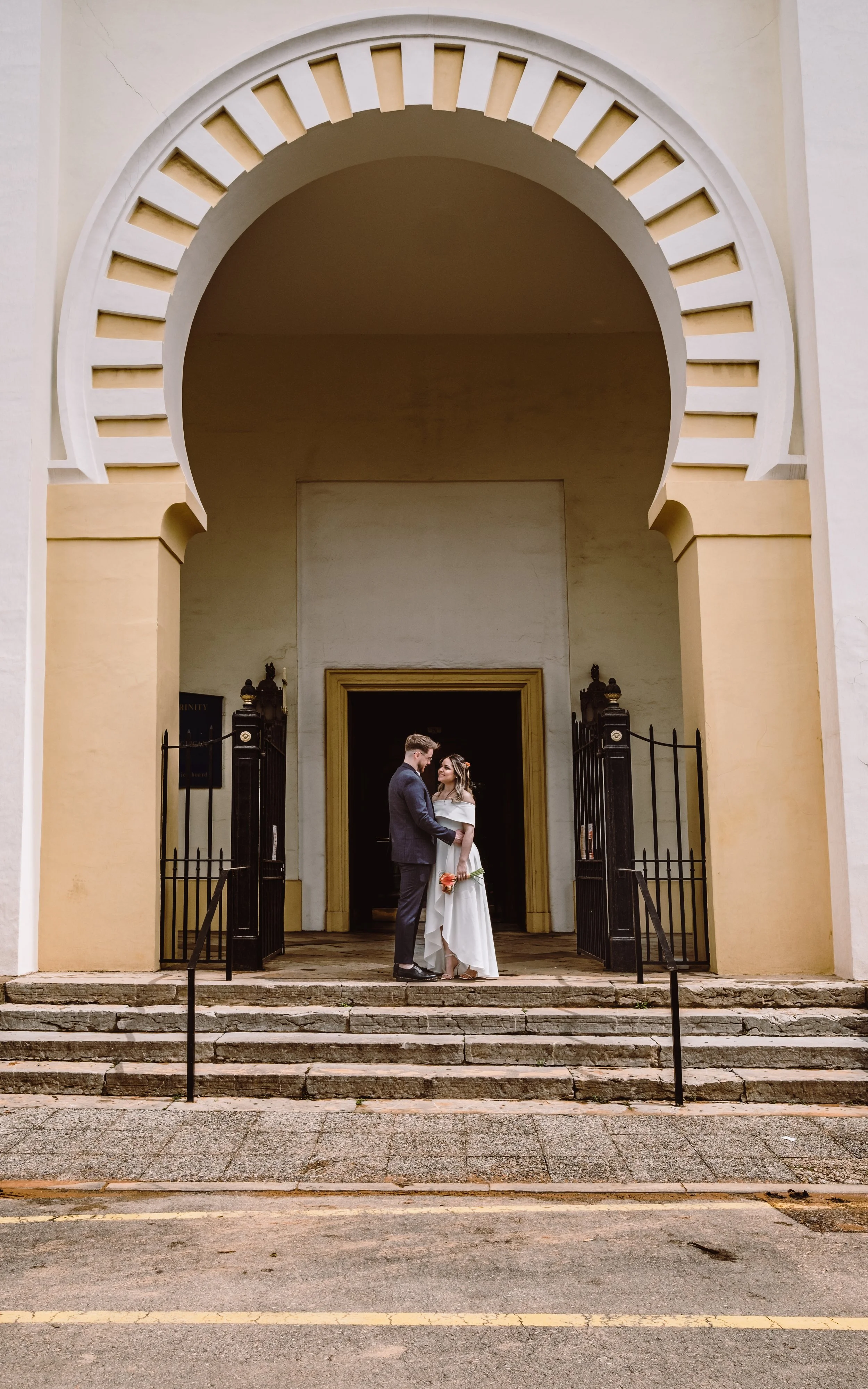 A bride and groom standing close together in front of a large, arched entrance with stairs, black iron gates, and yellow columns, outside a historic building.