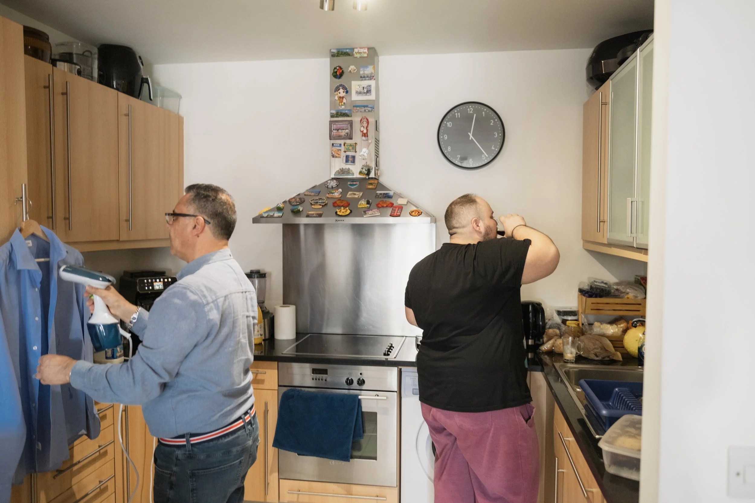 Two men standing and working in a kitchen with wooden cabinets. One man is holding a spray bottle, and the other is adjusting something on the wall. The kitchen has various magnets on a vertical surface, a wall clock, and appliances on the counters.