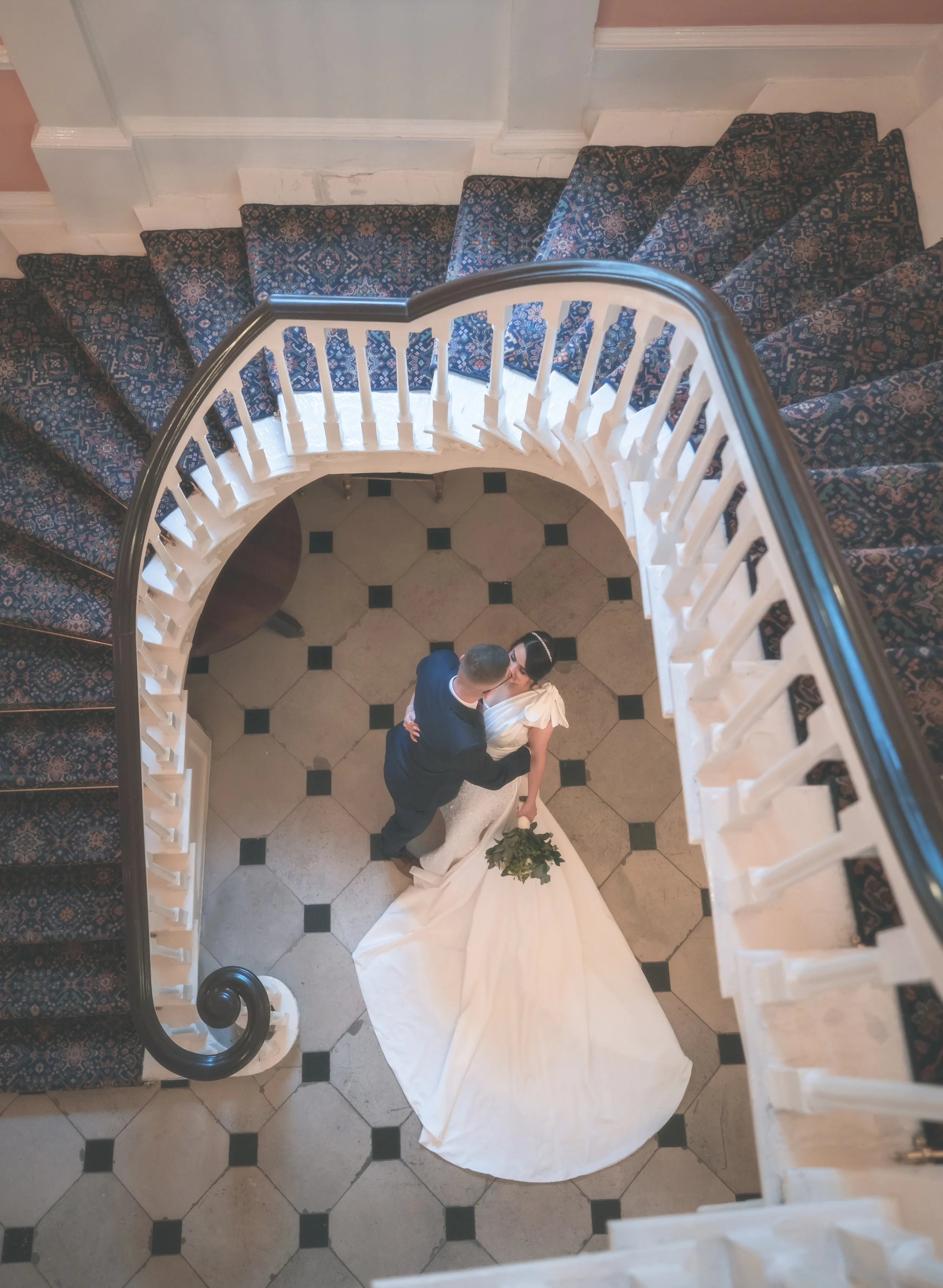 A bride and groom sharing a dance on a checkered tile floor at their wedding, viewed from above at the top of a curved staircase with a patterned carpet and white railing.
