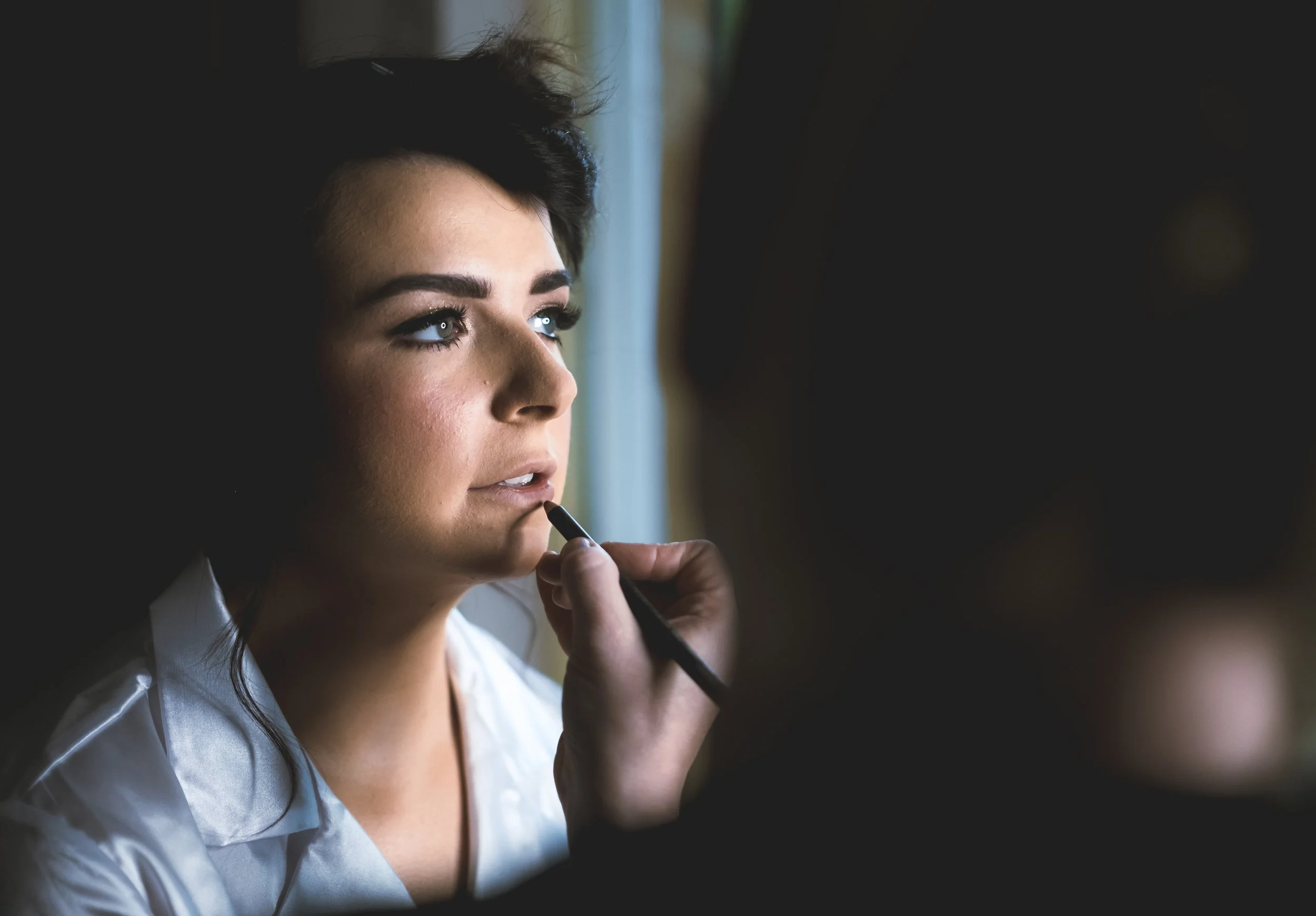 A woman with dark hair and makeup looking out a window while applying lipstick with a brush.