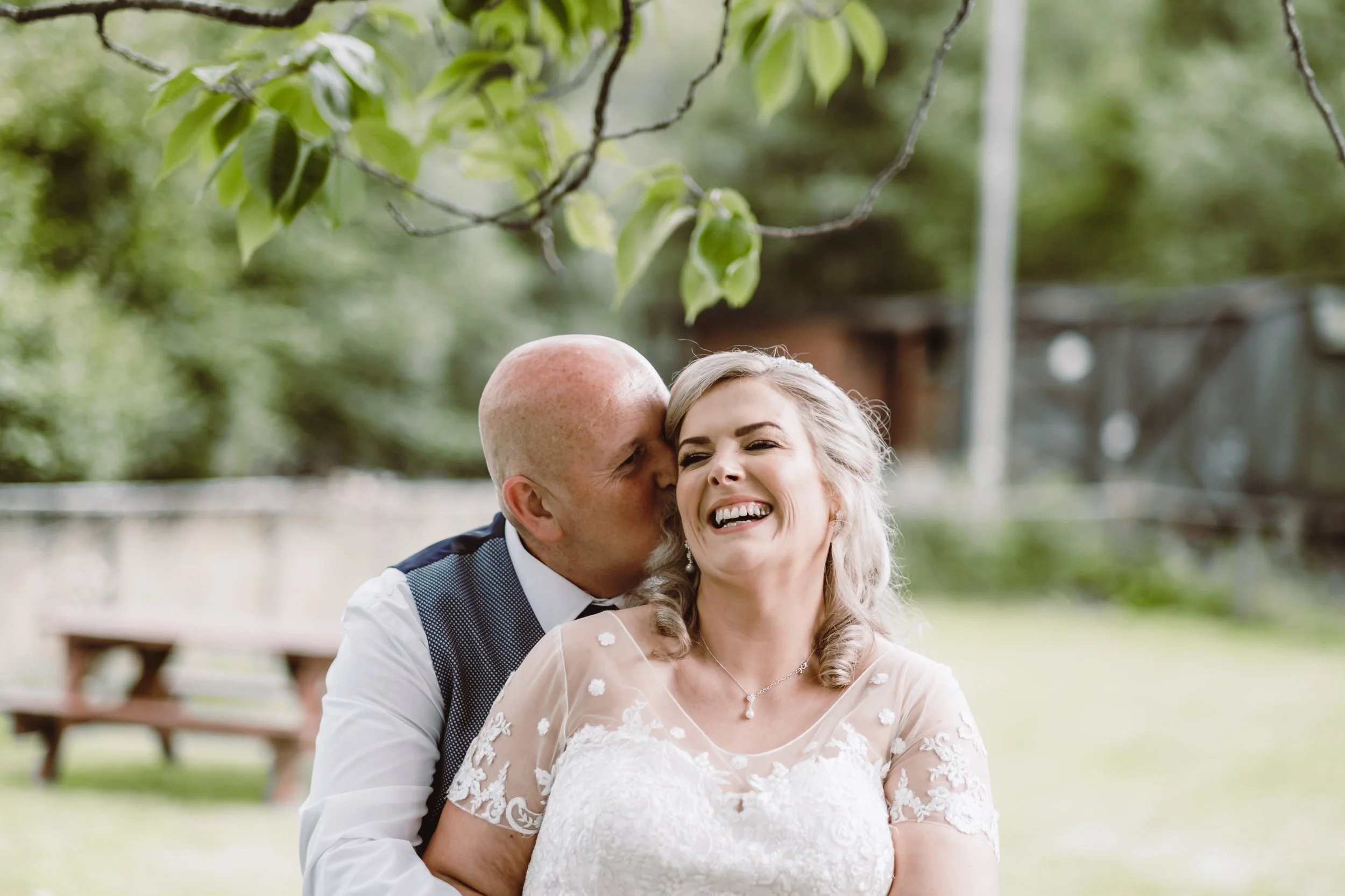 A couple on their wedding day, the groom with a bald head and the bride with blonde hair, smiling and embracing outdoors under a leafy tree.