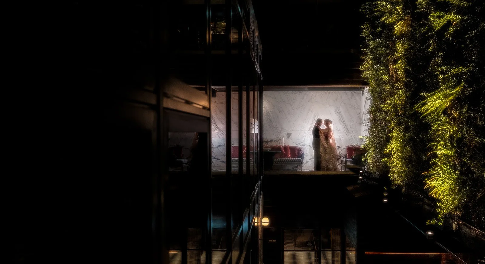 A couple standing close together in a warmly lit room with a marble wall, viewed through a glass window with a black metal frame. The room has furniture with red pillows and a lush green vertical garden illuminates the right side of the scene.