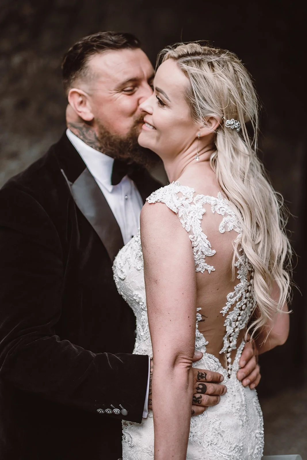 A bride and groom sharing a kiss on their wedding day. The bride is wearing a lace wedding dress with open back and long blonde wavy hair, while the groom is in a black tuxedo with a white shirt and black bow tie.