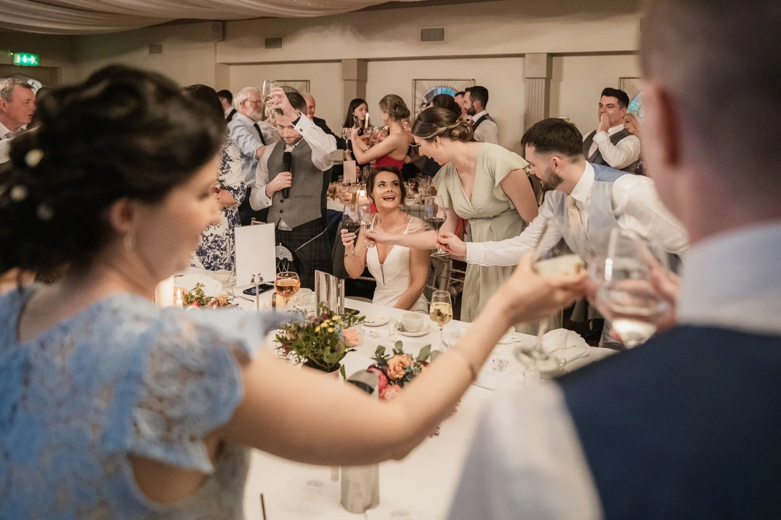 Guests at a wedding reception raising glasses in a toast, with a bride and groom sitting at a table, surrounded by friends and family, in a warmly decorated venue.