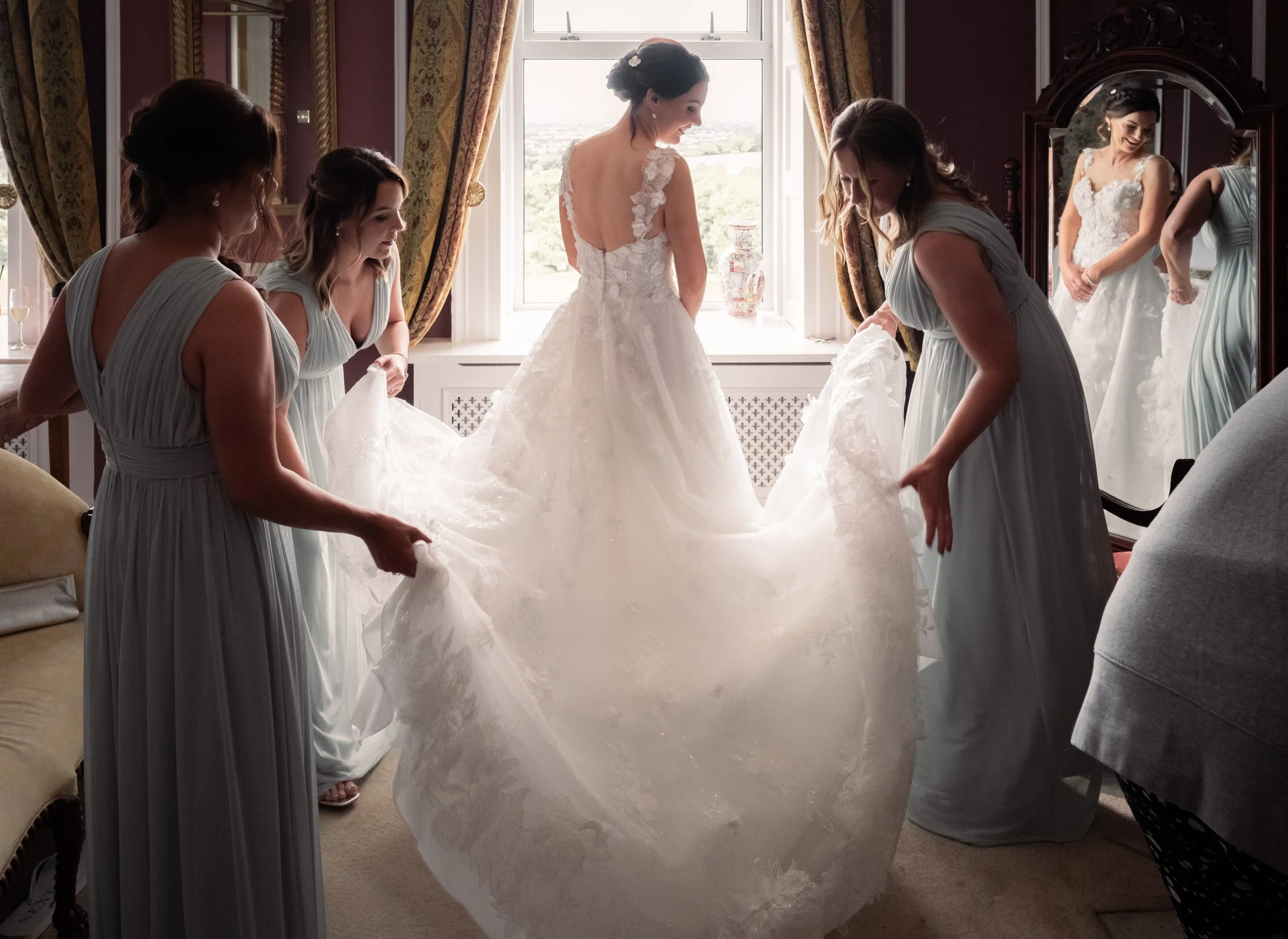 Bride in a wedding dress with lace details smiling at herself in a mirror, while three bridesmaids in matching light blue dresses assist her, in a room with large window, gold patterned curtains, and a vintage mirror.