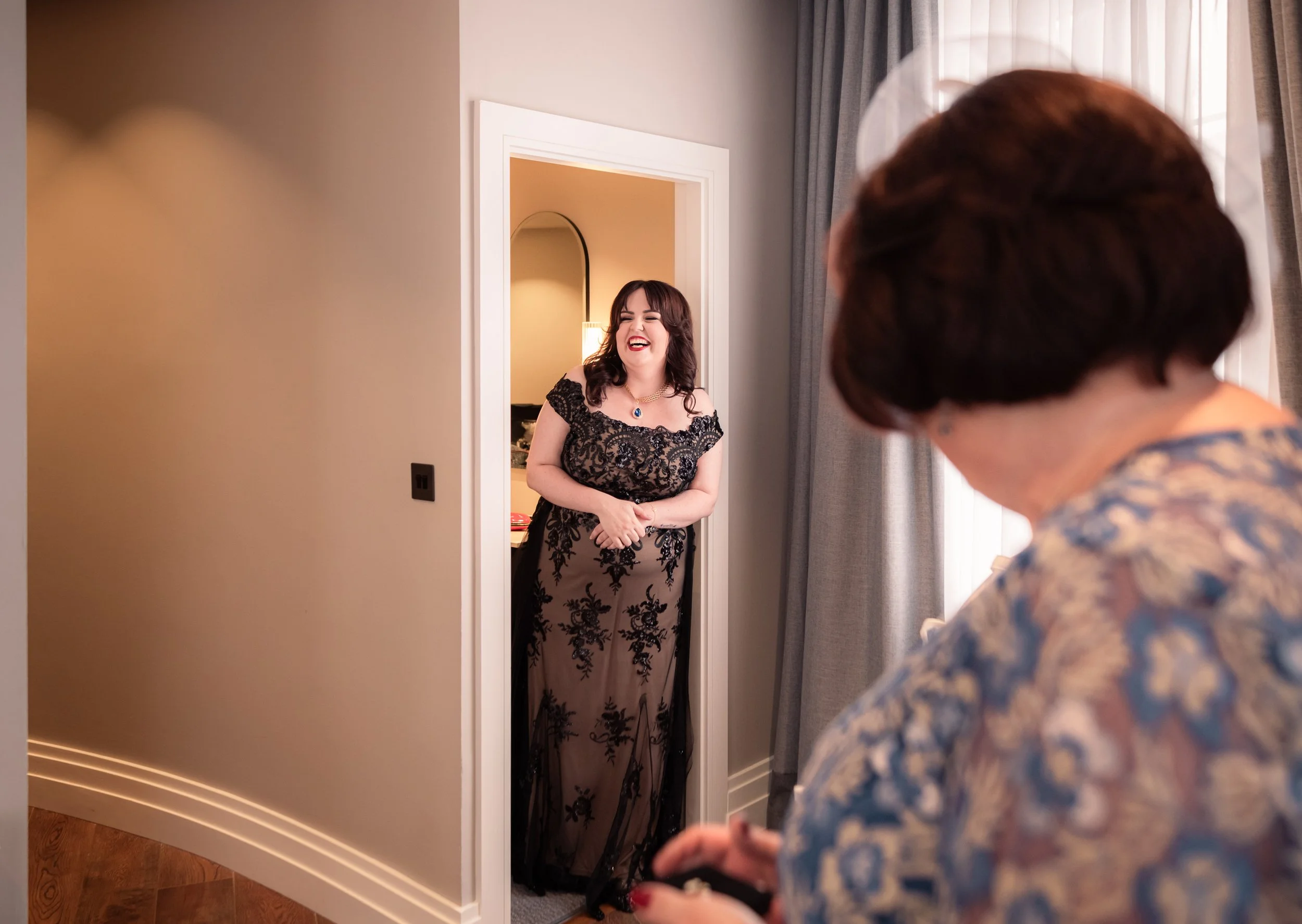 A woman in a black lace dress standing in a doorway, smiling, with another person in the foreground.