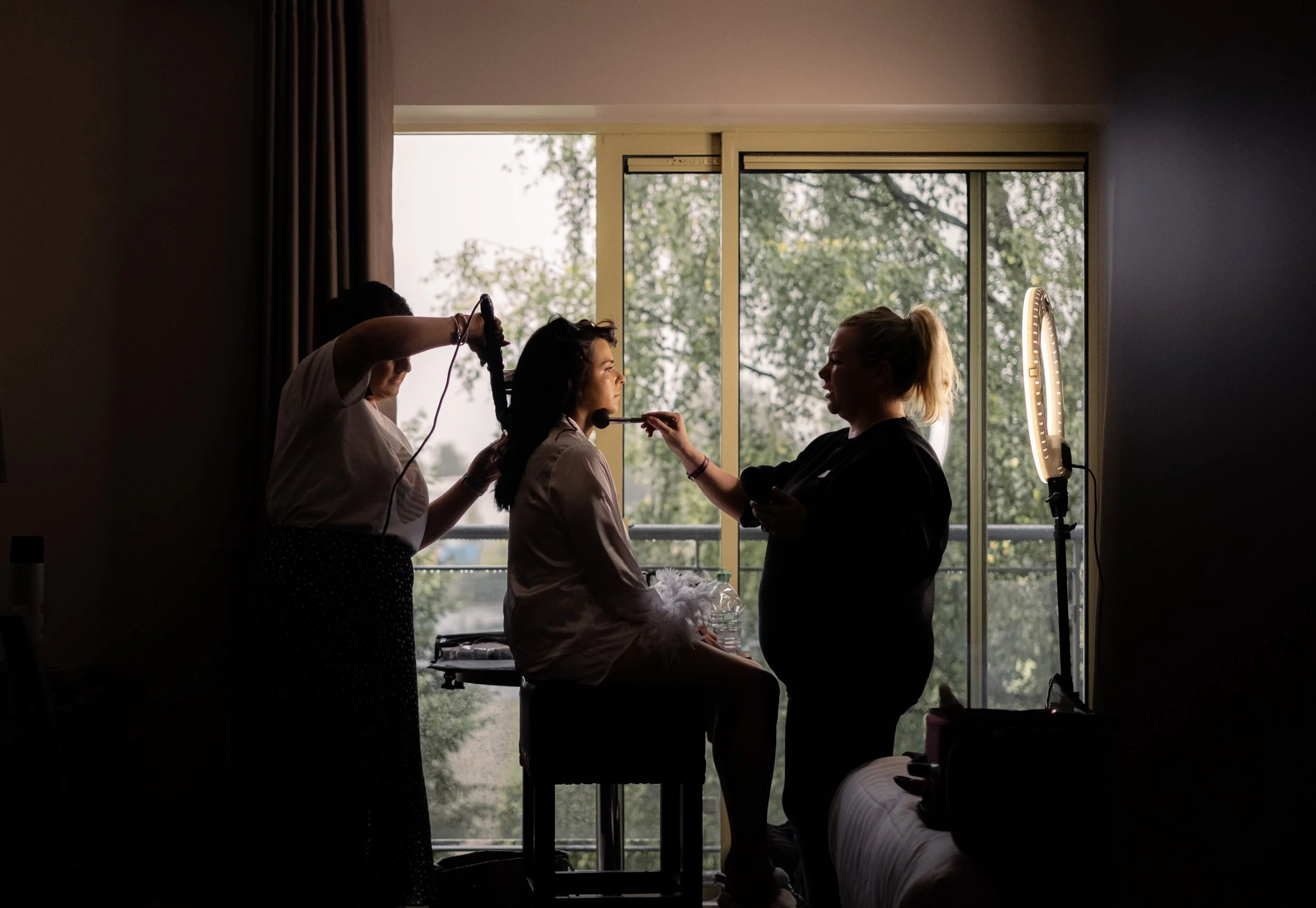 A woman sitting on a bed while makeup artists work on her face, with a large window showing trees outside and professional lighting equipment nearby.
