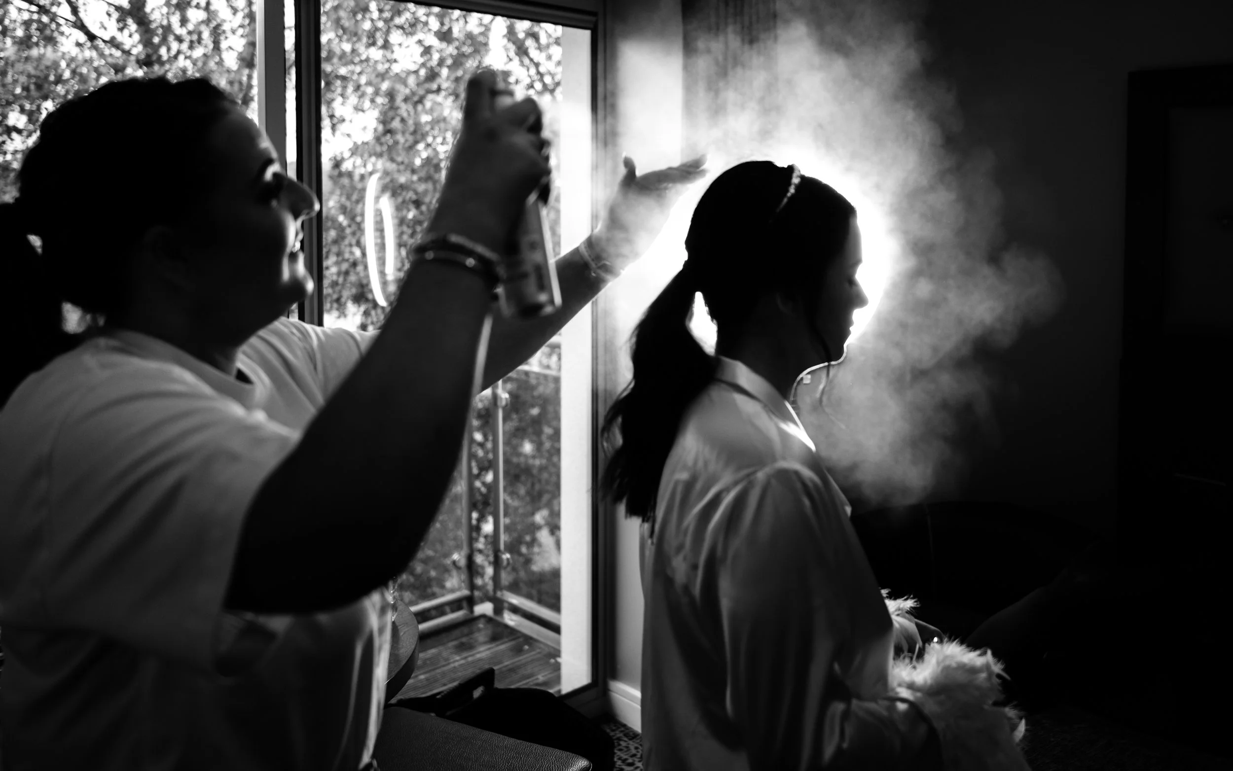 A woman applying hairstyling spray to another woman's hair in a dimly lit room with sunlight coming through a window.