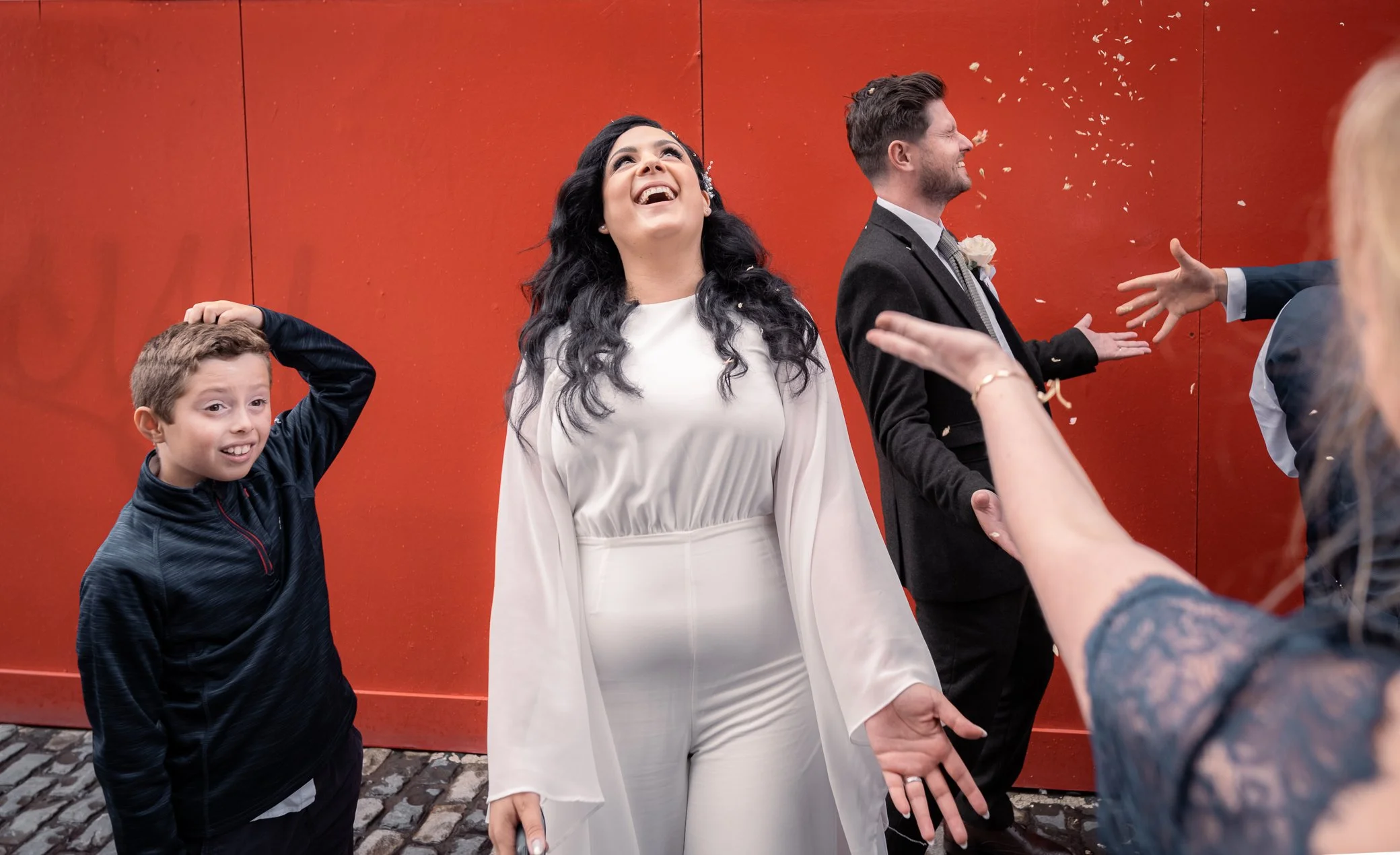 A woman in a white outfit celebrating with a joyful expression, surrounded by children and adults at a celebration against a red background.
