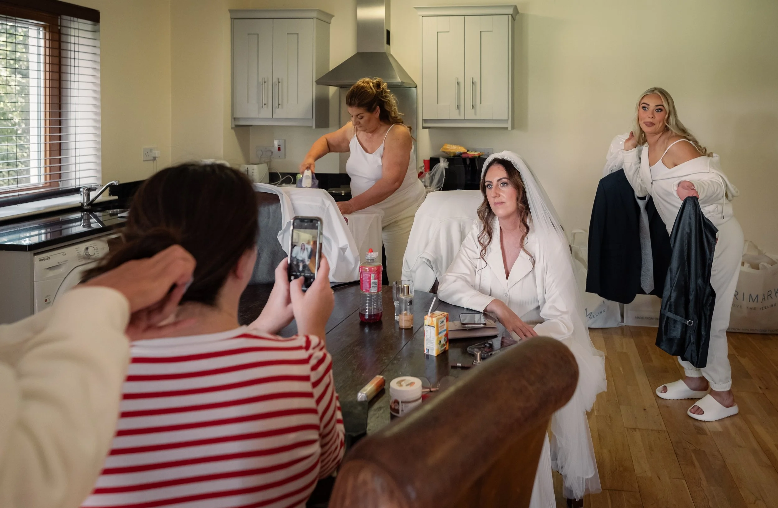 Women preparing for a wedding in a kitchen; one woman is ironing a white dress, another is wearing a white dress with a veil, and a woman on the right is adjusting her outfit; people are taking photos and sitting at a table with drinks and snacks.