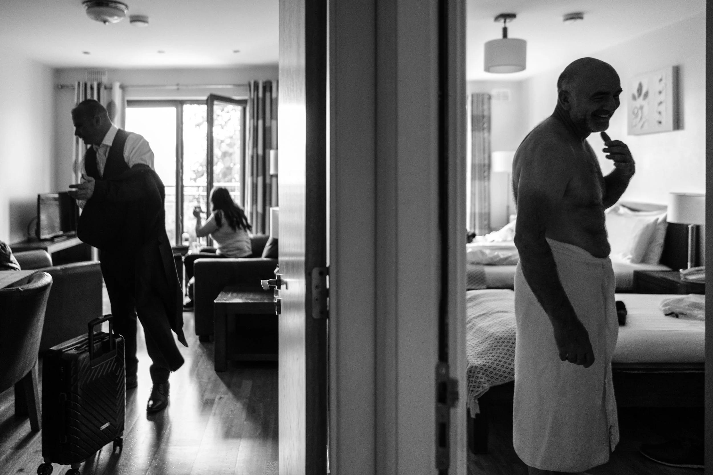 A black-and-white photo showing two scenes separated by a door frame within a room. On the left, a man in a suit is standing, looking at his phone, with a woman sitting on a sofa in the background near a window. On the right, a man is standing shirtl