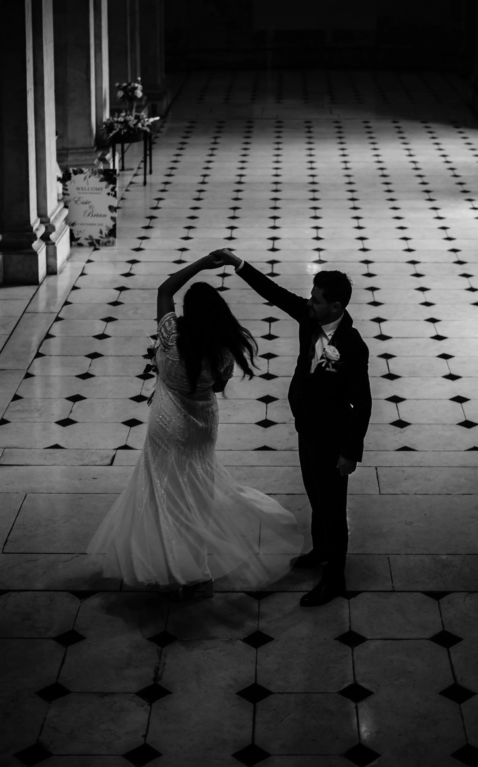 A black and white photo of a bride and groom dancing in a large hall with a patterned tile floor. The bride is in a long, flowing wedding dress, and the groom is in a dark suit. The bride's hair is long and wavy, and the groom has short hair. There a