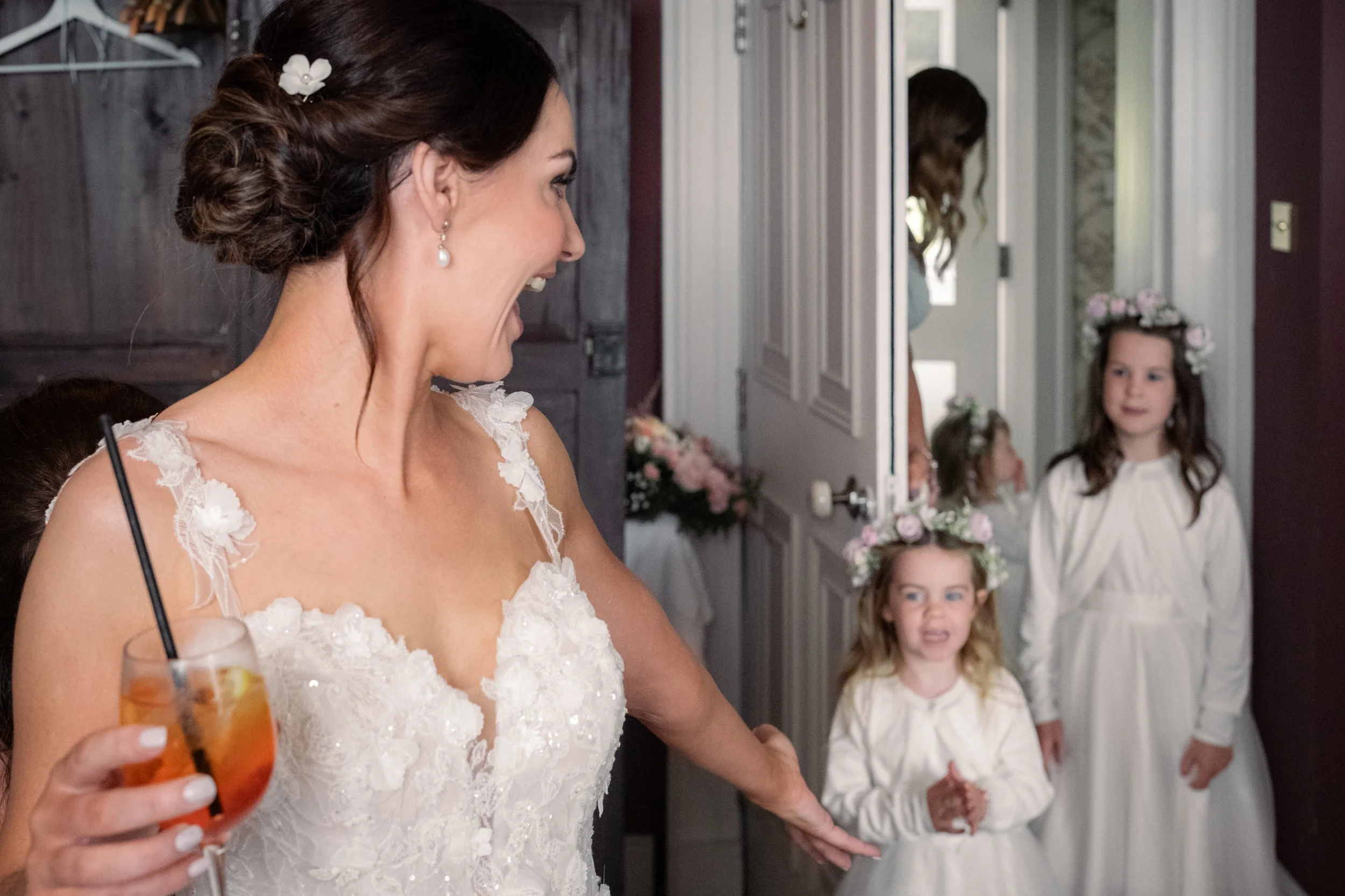 Bride in a lace dress joyfully gestures toward two young flower girls wearing floral crowns and white dresses, entering a room. The bride holds a drink with a straw.