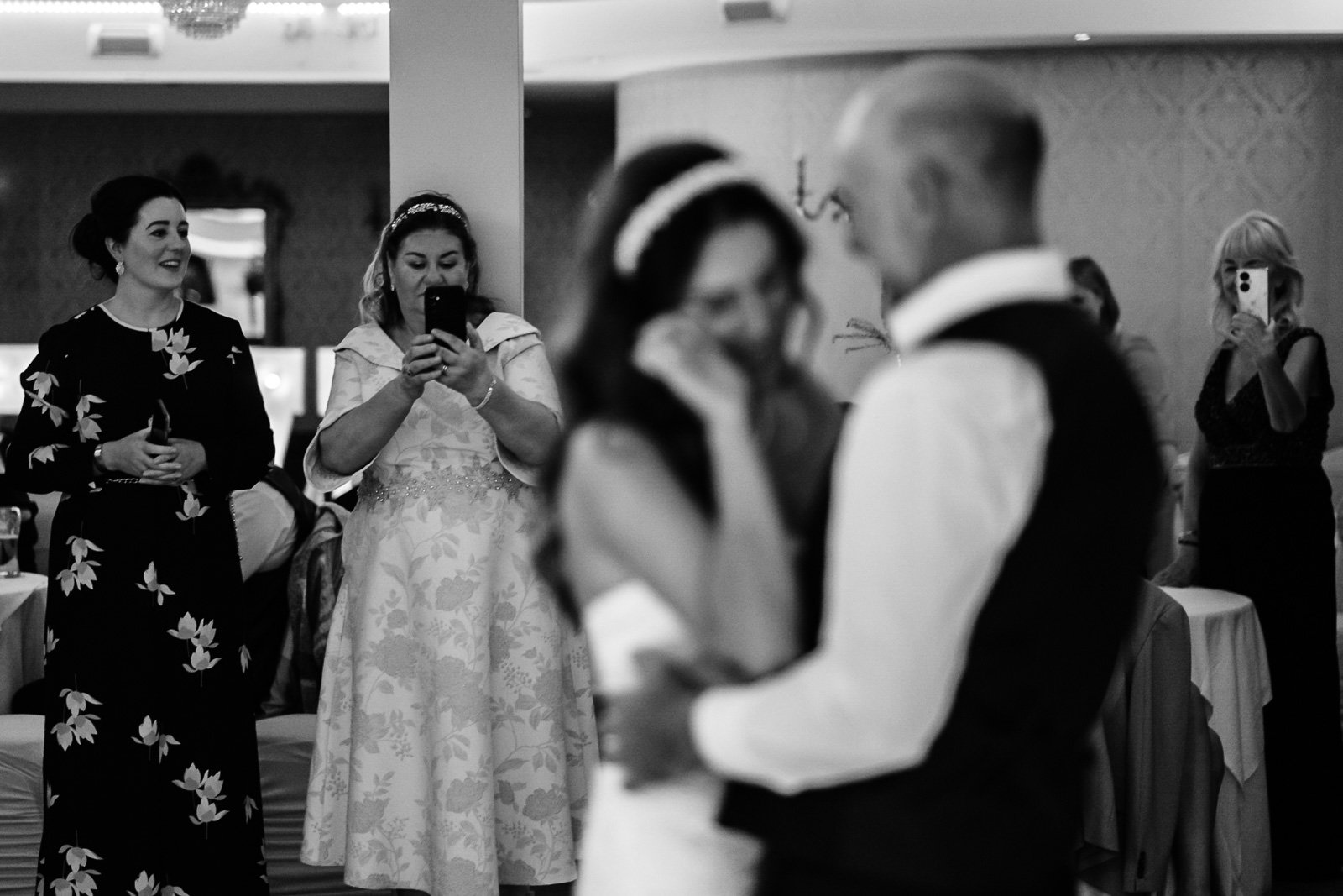 A black-and-white photo of a wedding reception showing a bride and groom dancing, with several women in the background taking pictures and watching.