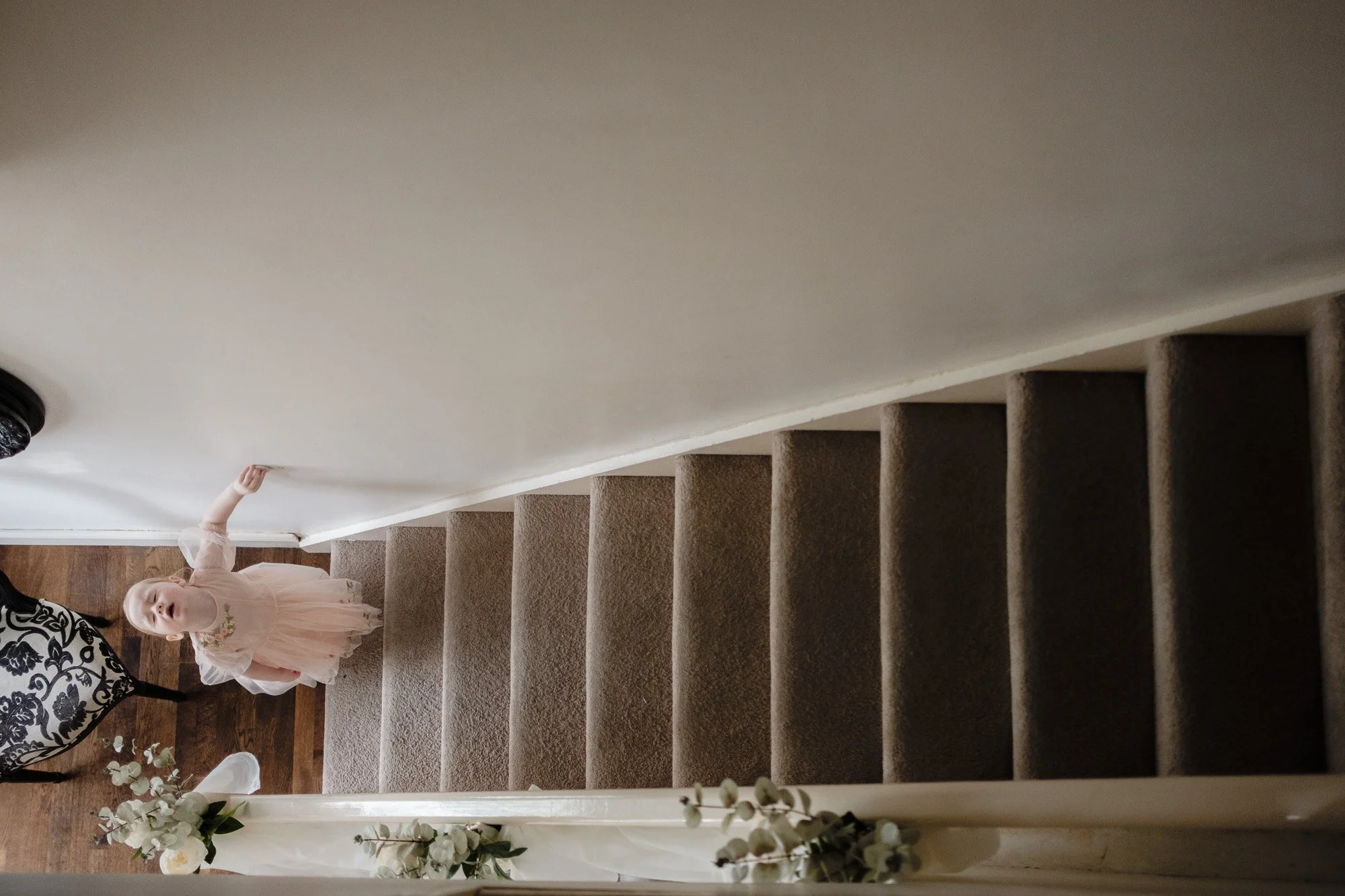 Top view of a young child in a pink dress standing at the bottom of a carpeted staircase, looking up. The stairs are decorated with white flowers.