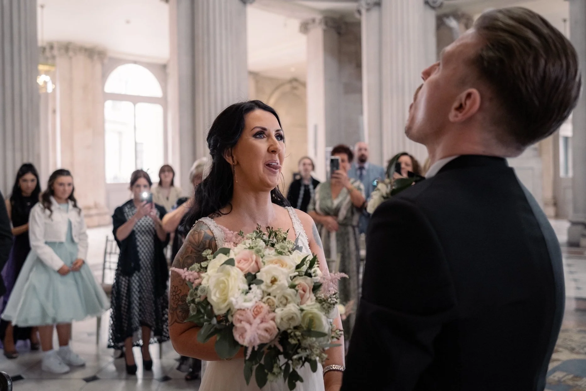 A bride and groom exchanging vows during a wedding ceremony inside a grand hall with tall columns. The bride is holding a bouquet of flowers and has dark hair, while the groom has light-colored hair and is dressed in a black suit. Guests are watching