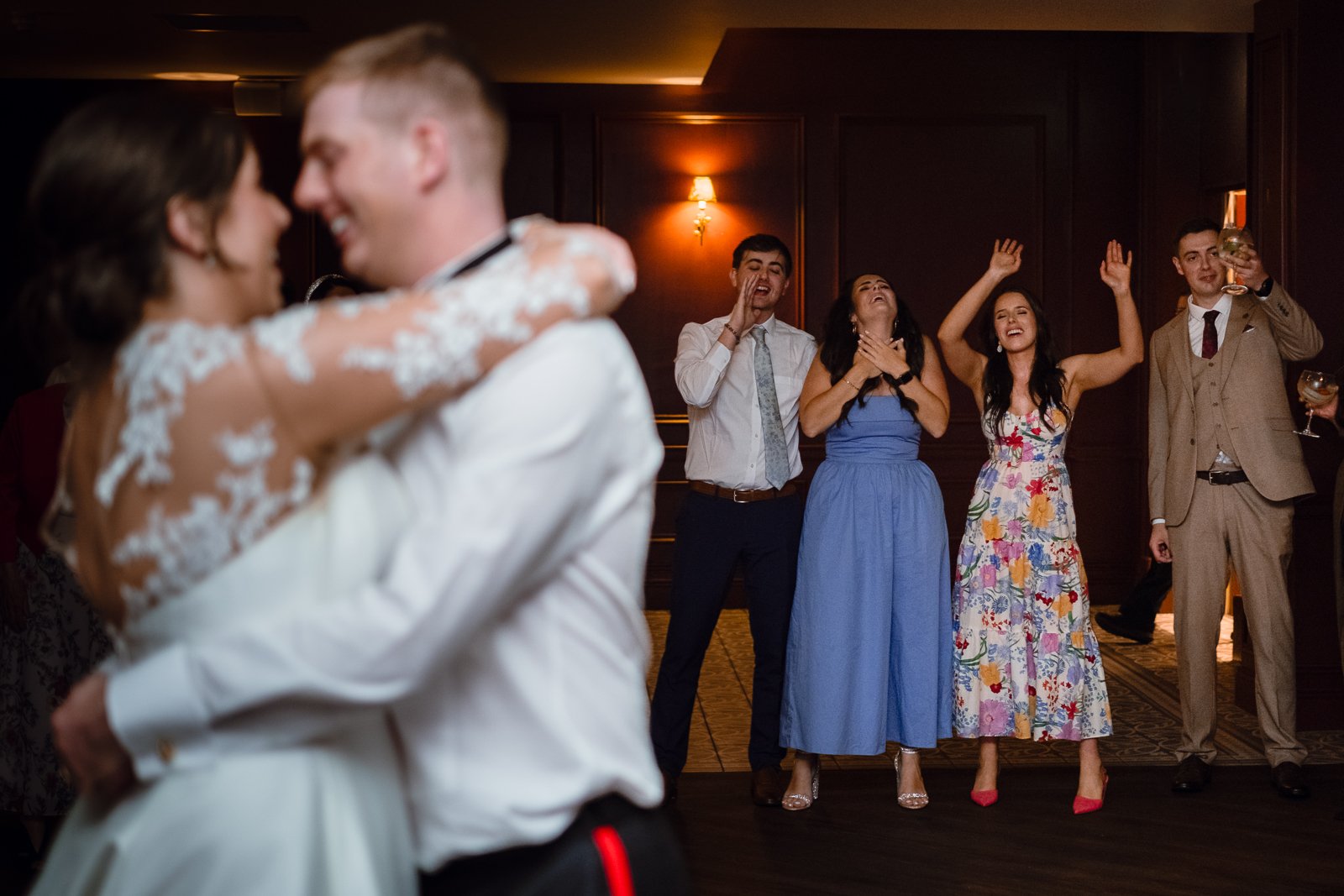 Couple dancing at a wedding while friends on the dance floor dance and celebrate in the background.