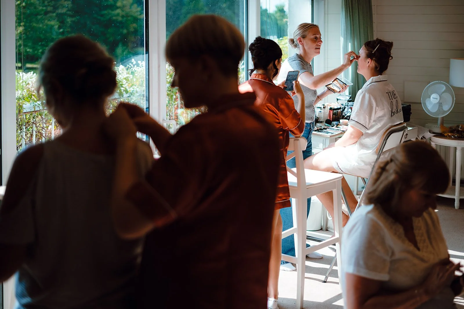 A woman applying makeup to a seated bride, with several women preparing and taking photos nearby inside a well-lit room with large windows and a view of greenery outside.