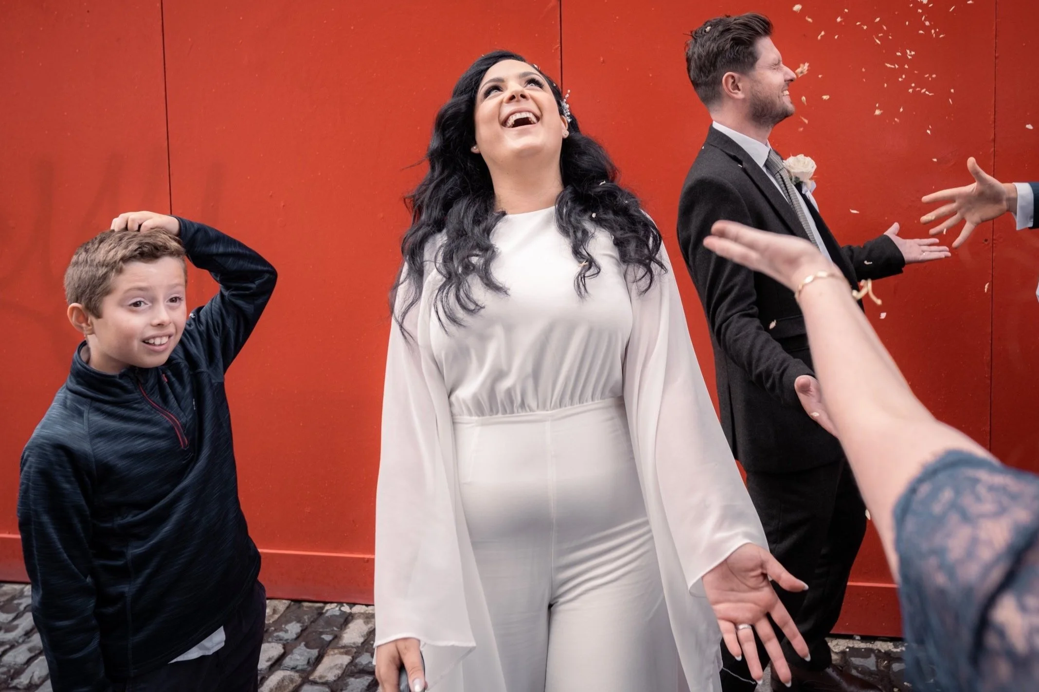 A woman in white celebrating at a wedding, surrounded by family and friends, with a red wall in the background.