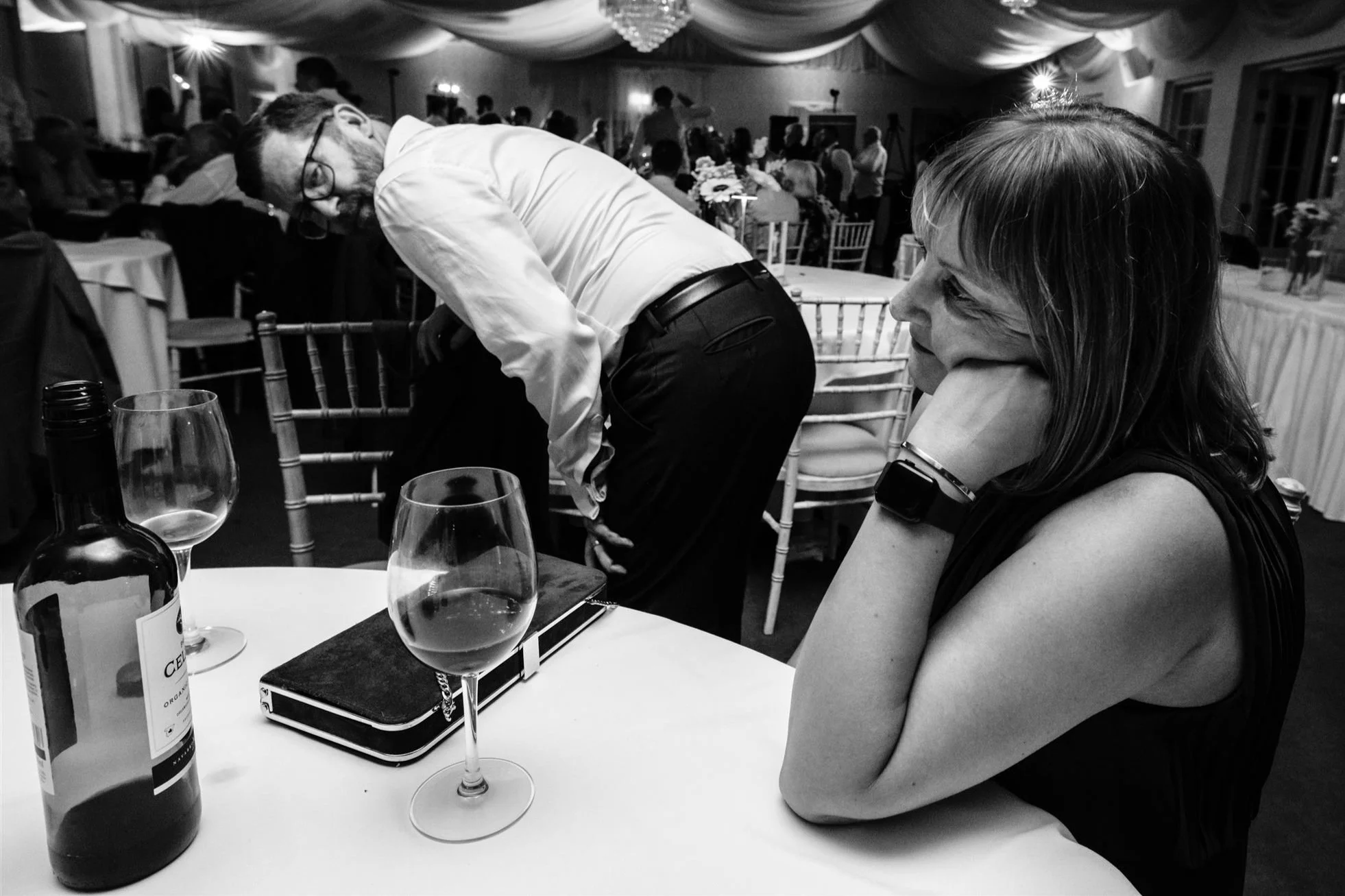 A woman sitting at a table with her head resting on her hand, watching a man bending over in a formal setting with other seated guests, decorated tables, and a ceiling with draped fabric, indicative of a party or wedding reception.