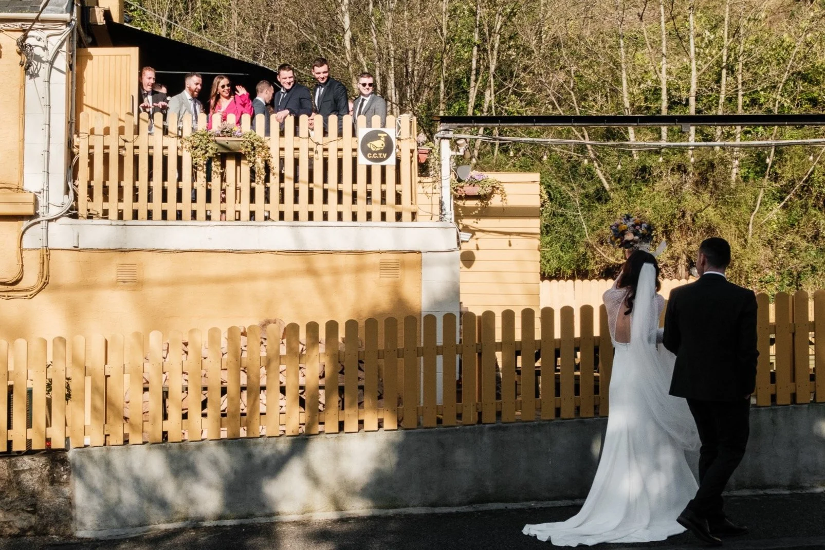 A bride in a white wedding dress and veil walking with a groom in a black suit, facing away from the camera, on a street with a beige fence. On the upper level, several men and women dressed formally are gathered, watching and smiling. The scene appe