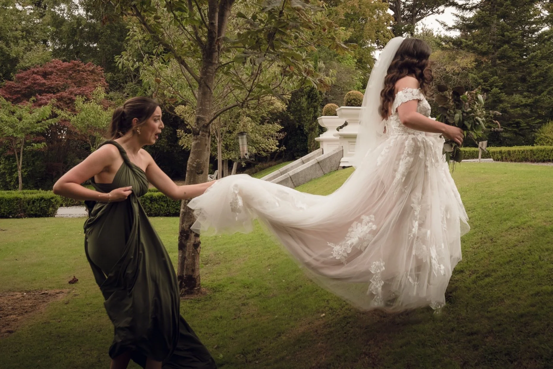A bride in a white wedding dress is being pulled by a woman in a dark green gown. The bride is holding a bouquet, and the scene is outdoors with trees and a grassy area.