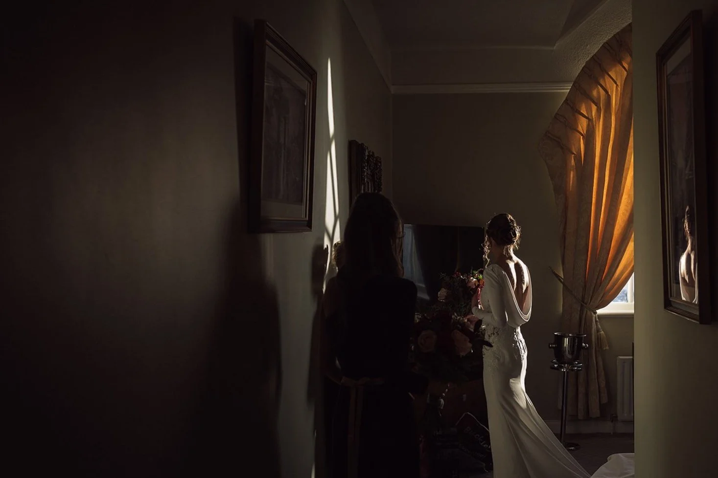 A bride in a white wedding dress standing near a window with golden curtains, holding a bouquet, while a woman, possibly a wedding coordinator or friend, stands nearby in a dimly lit room.