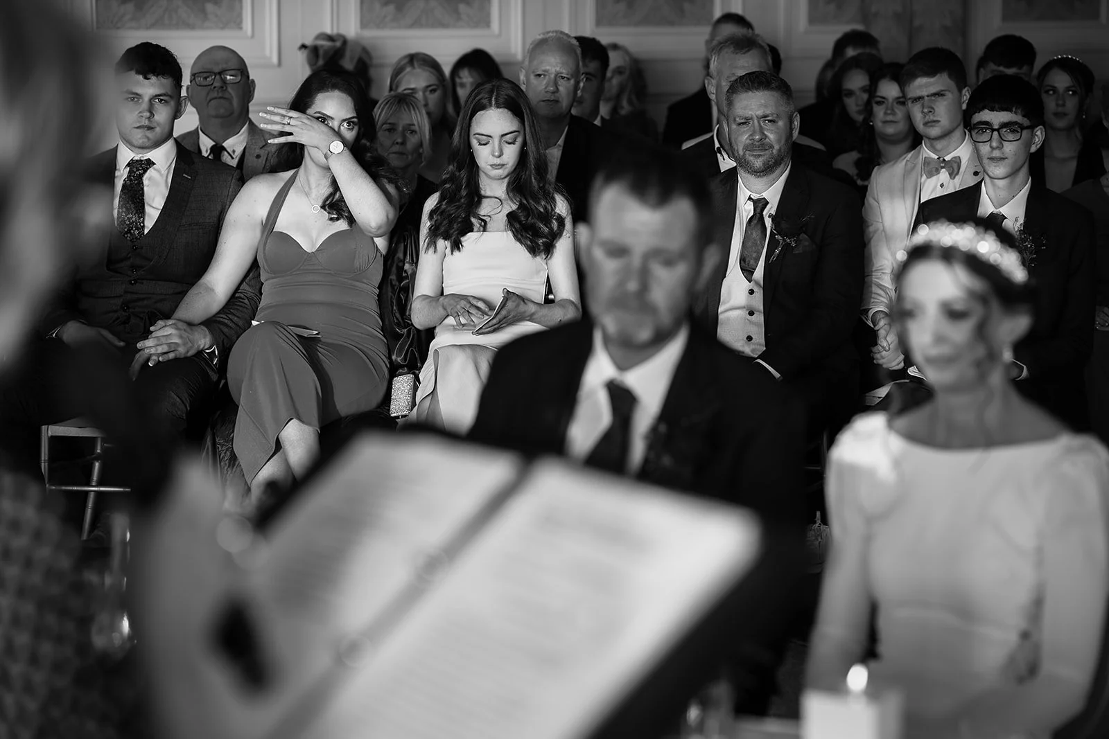 A black-and-white photo of wedding guests seated in an indoor ceremony, with some appearing emotional or focused, and the officiant reading from a book at the front.