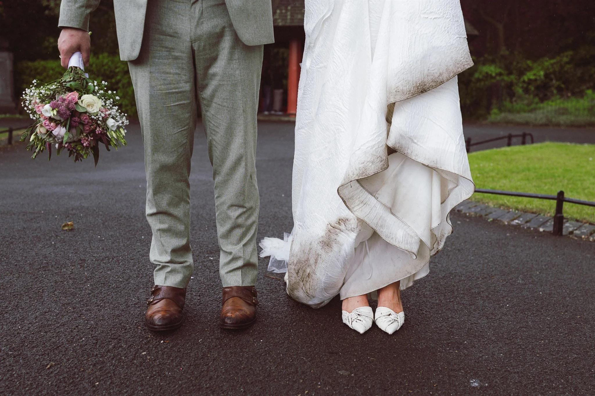 A couple standing outdoors, the groom holding a bouquet of flowers, with the bride wearing a white wedding dress and shoes.