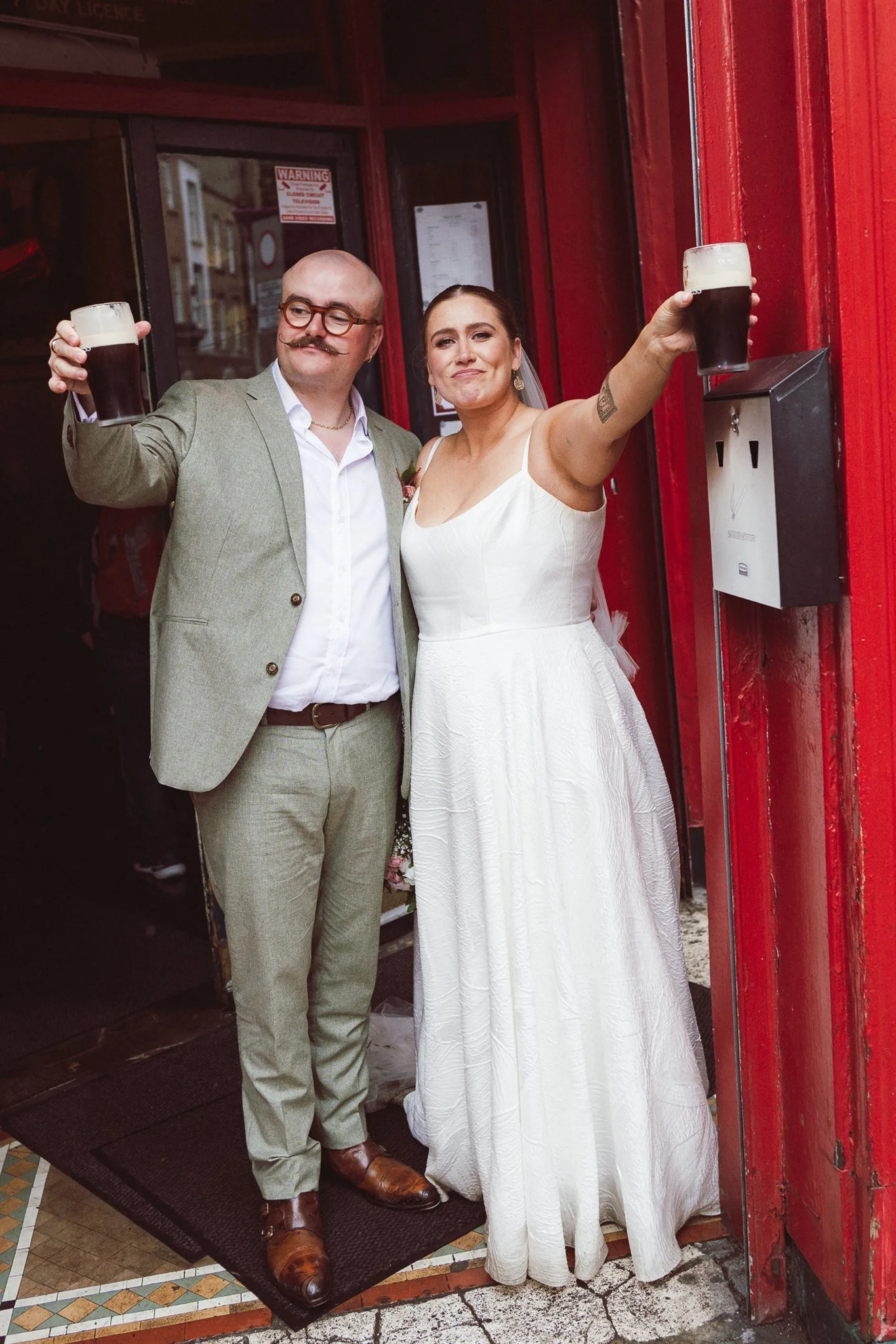 A newlywed couple in wedding attire celebrating outside a venue, holding glasses of dark beer with foamy heads.