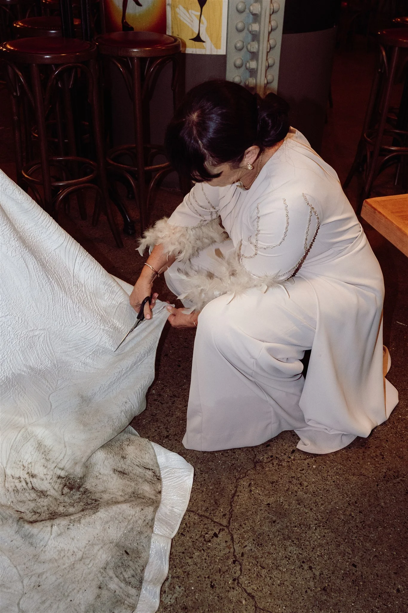 A woman in a long, elegant white dress kneels on the floor, cutting a white wedding dress with scissors in a dimly lit room.