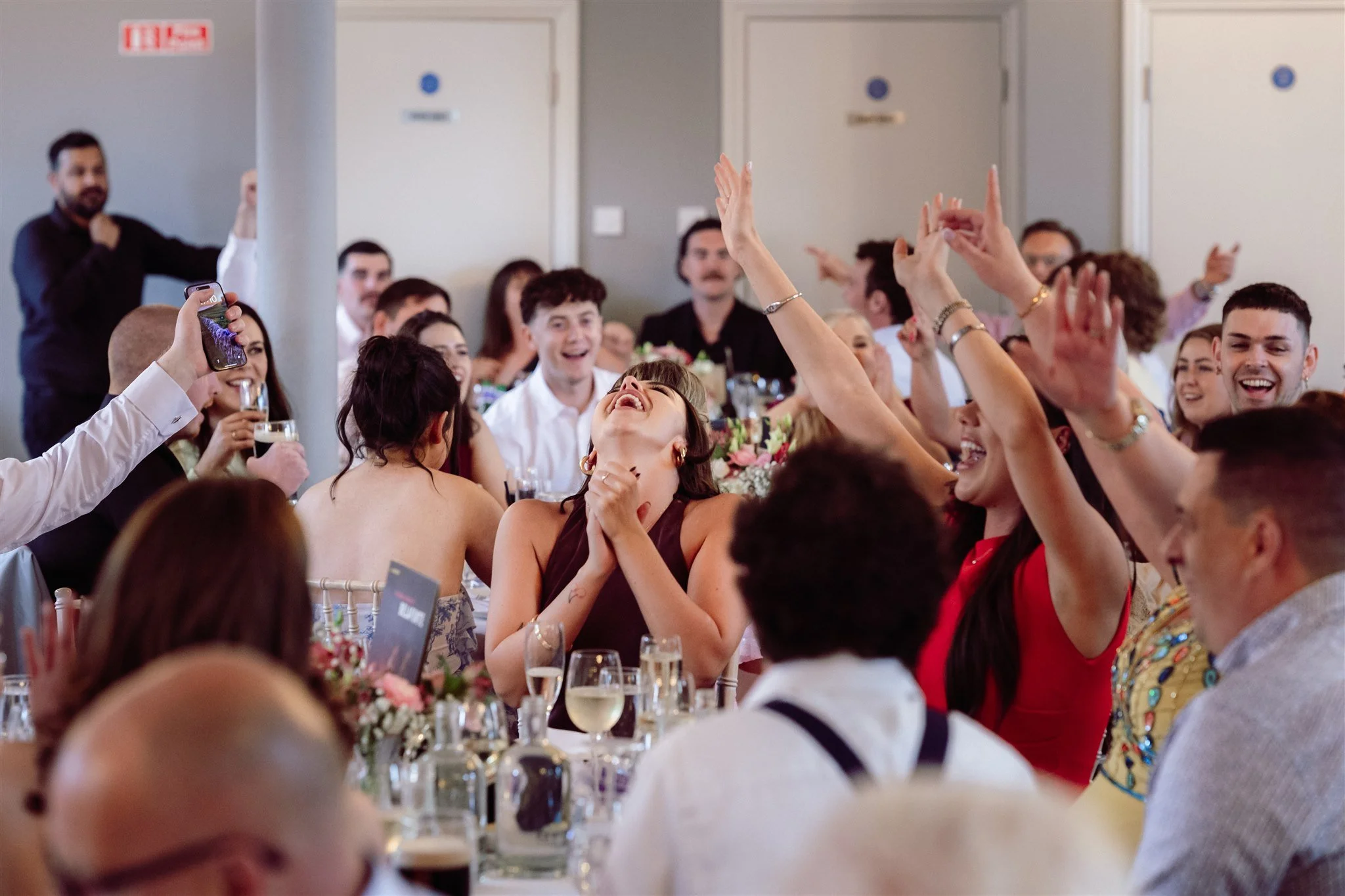 People celebrating at a banquet, some with drinks, with one woman in the center laughing and pointing upwards, in a lively and joyful gathering.