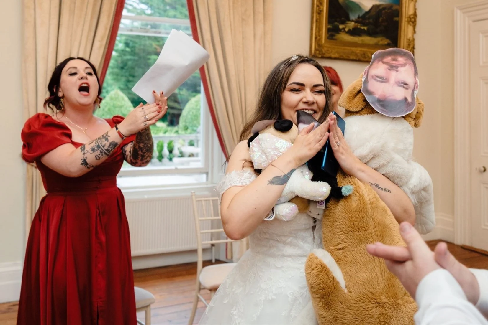 A bride in a white wedding dress is holding a large stuffed dog toy with a face mask on its head. She is smiling and appears happy. A person or child's hand is pointing to her from the right. In the background, a woman in a red dress is holding a pap
