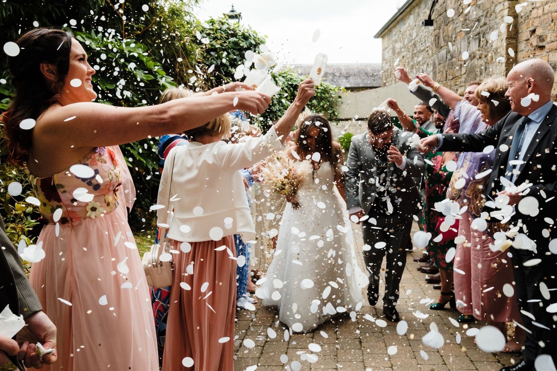 A bride and groom walking through a tunnel of friends and family throwing confetti at them during their wedding celebration outdoors.