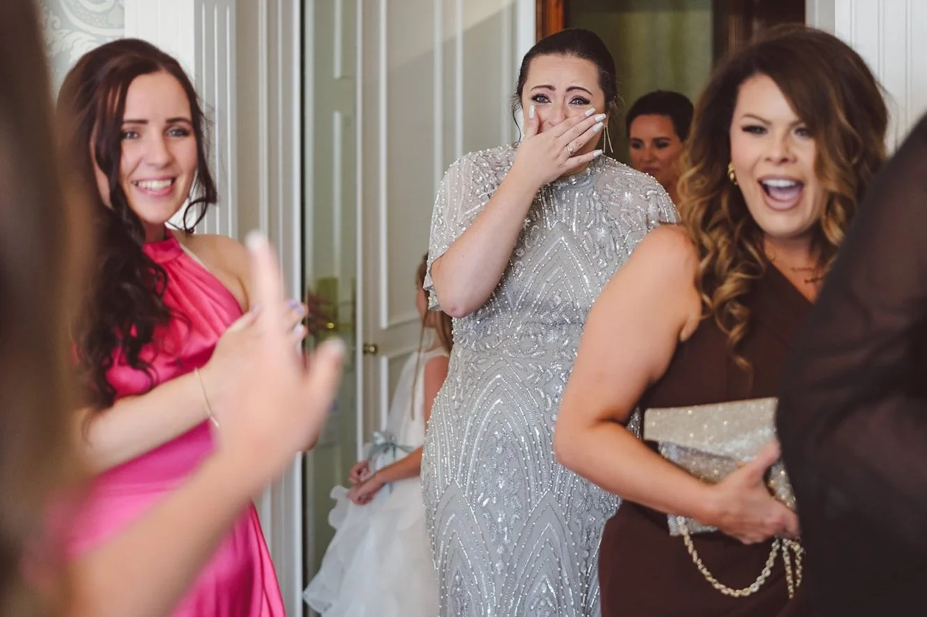 A group of women celebrating at a joyful event, possibly a wedding reception, with one woman covering her mouth in surprise and others smiling and laughing.