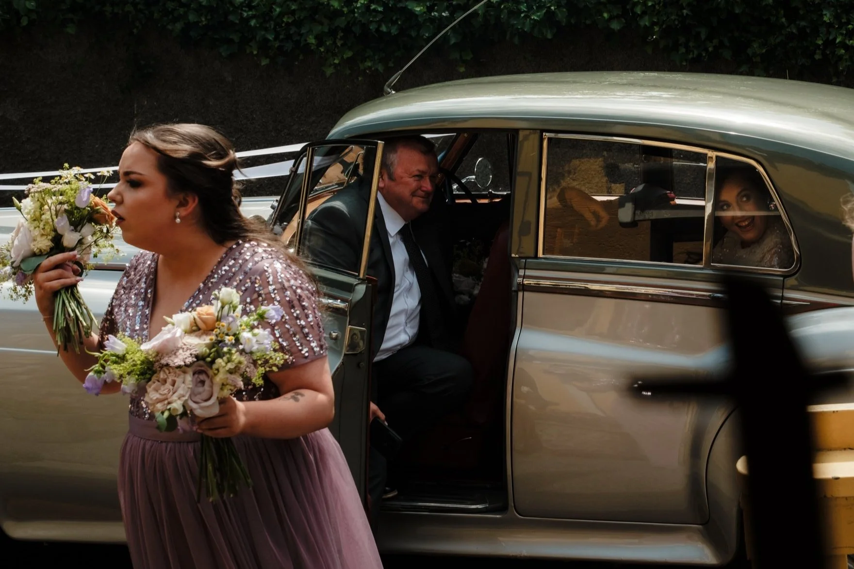 A woman in a glittery dress holding a bouquet of flowers walking past a vintage car, with a man and a woman inside the car, the man smiling and the woman smiling through the window at the back.