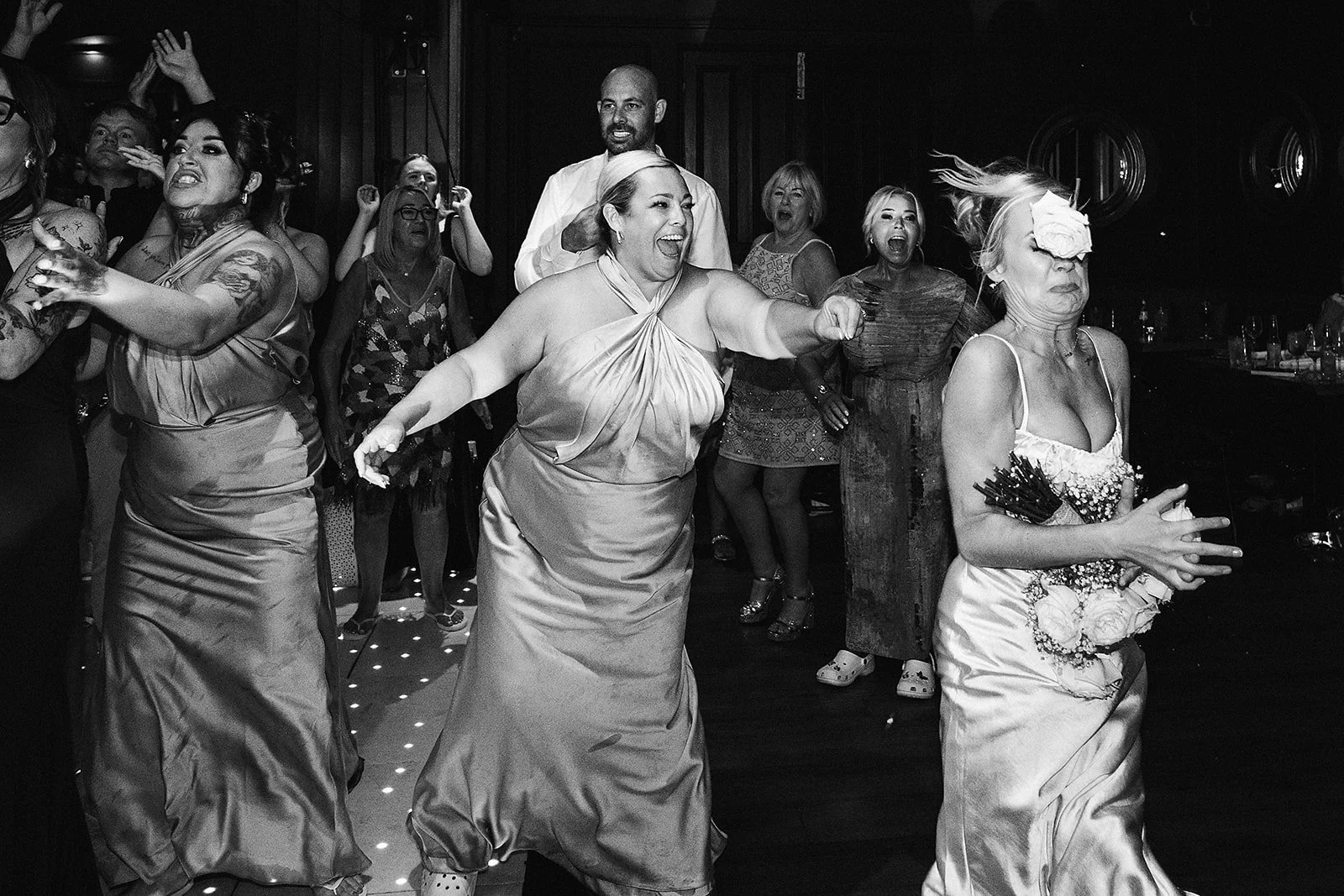 People at a lively event dancing and celebrating, with some women in elegant dresses and a man in a white shirt, in a dimly lit setting.