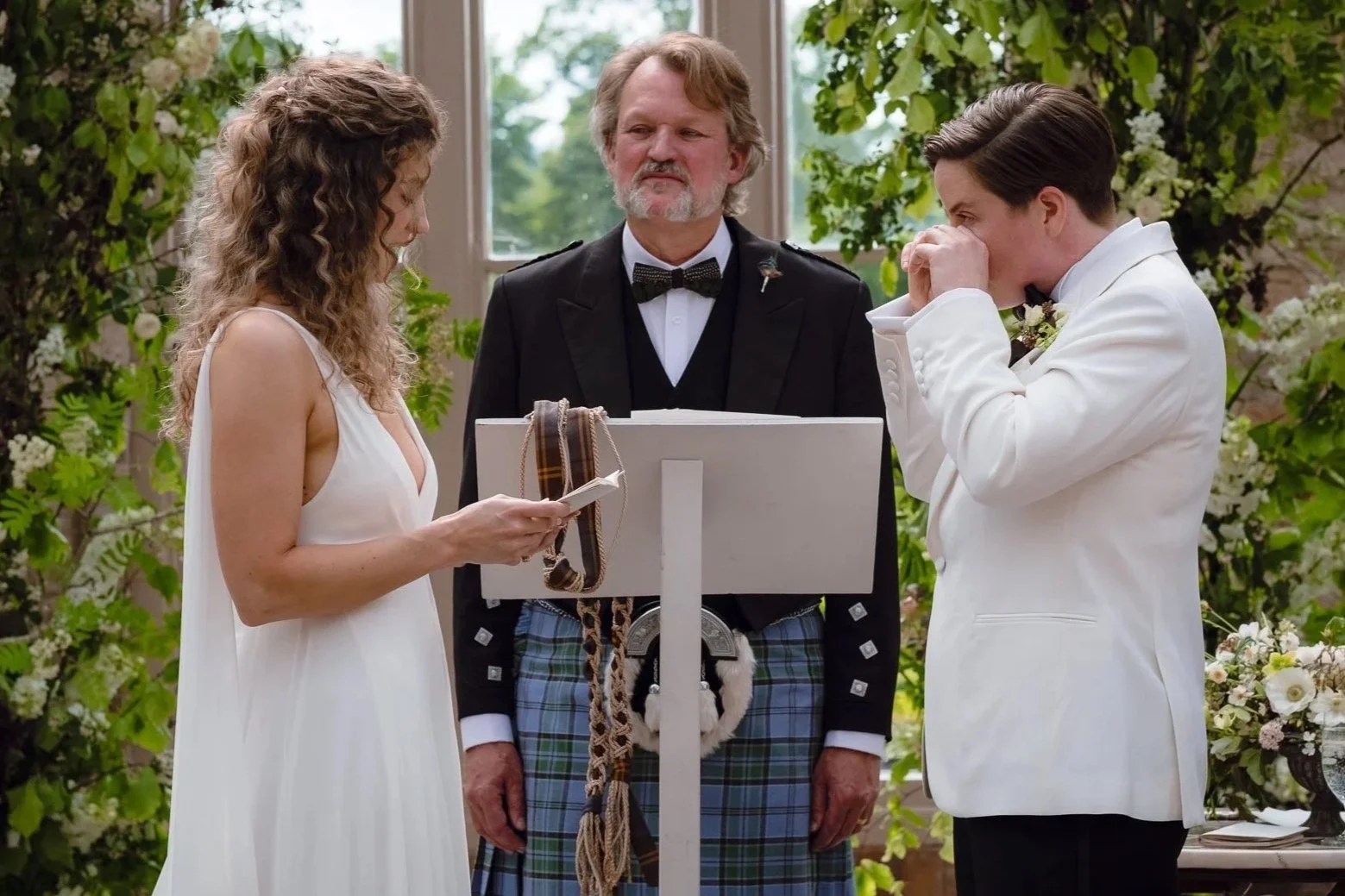 A couple getting married, standing before an officiant during a wedding ceremony in a lush, green setting. The bride is in a white dress, and the groom is in a white tuxedo. The officiant is dressed in a tuxedo with a kilt and is reading from a book 